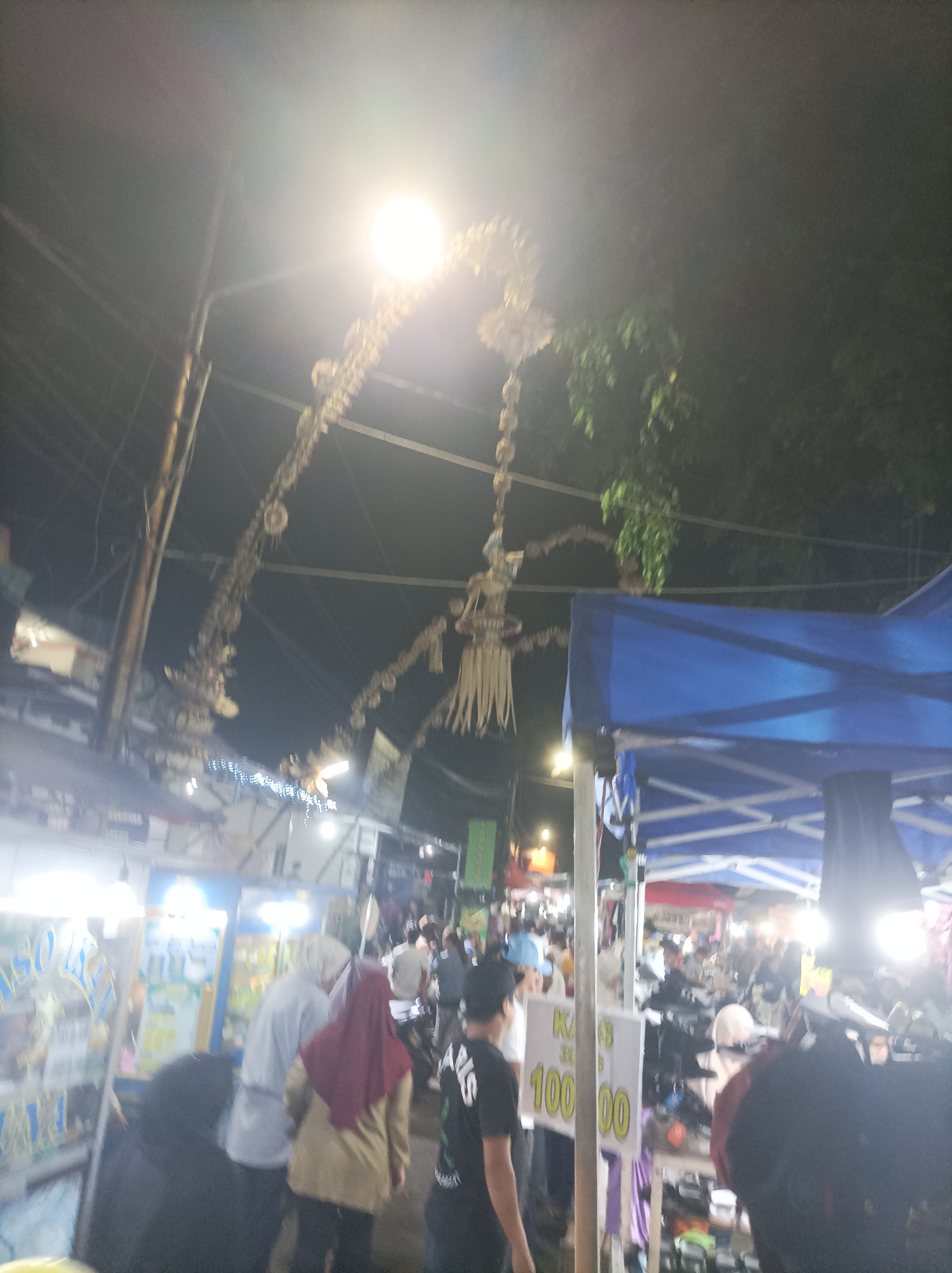 The image depicts a night market scene in Cirebon, Indonesia. In the foreground, several people are visible, some wearing hijabs, suggesting a predominantly Muslim population. One individual is wearing a black t-shirt with white lettering. There are stalls with merchandise, including what appear to be shoes and clothing. A blue tent is prominently featured. Decorative elements, possibly traditional decorations, hang from overhead structures, illuminated by bright lights. The overall atmosphere suggests a lively and bustling marketplace at night.