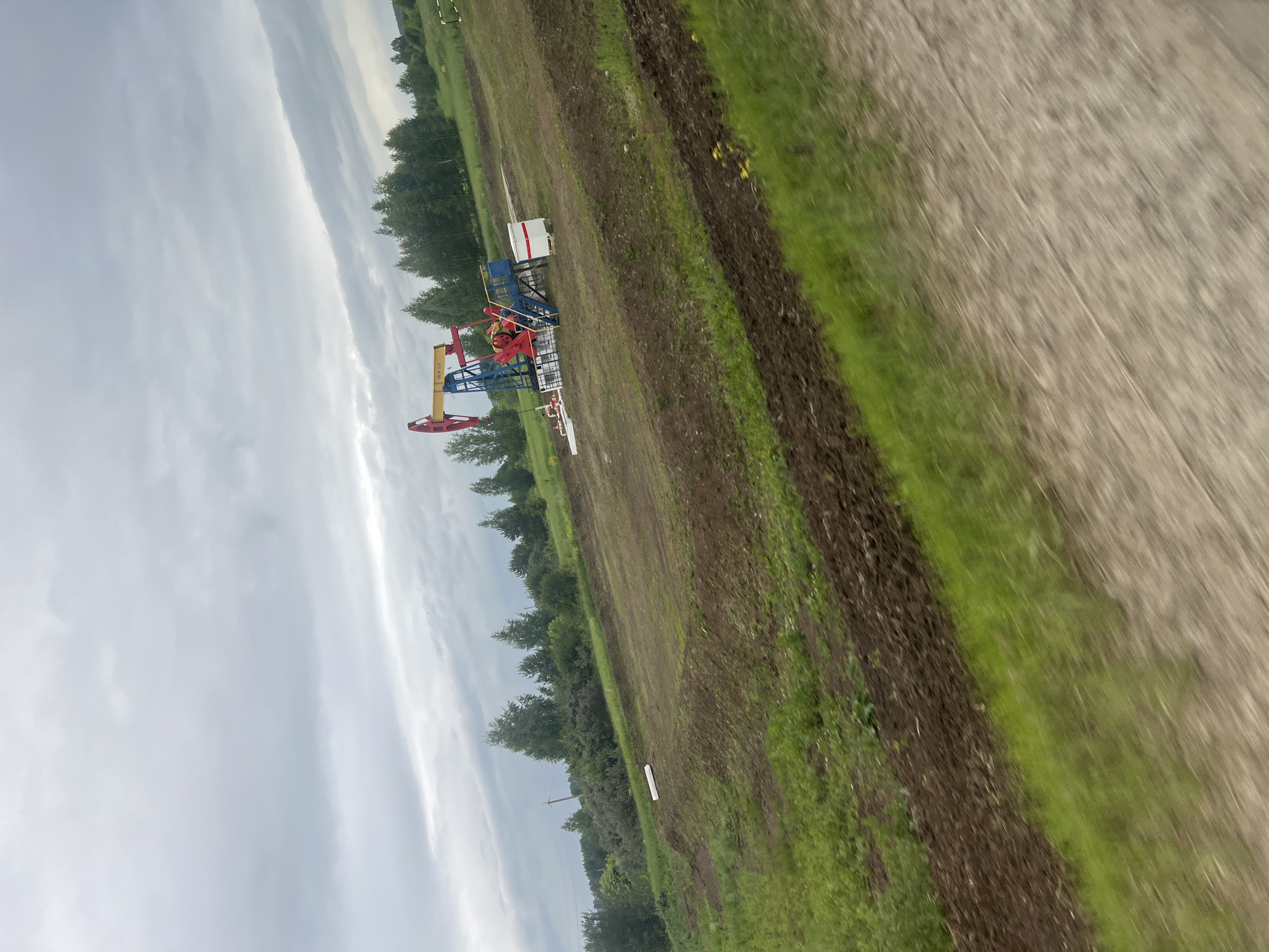 In Ustinovo, Russia, a pumpjack stands in a grassy, disturbed landscape. The pumpjack is primarily blue and red with a yellow beam. A small white structure with a red roof is positioned near the pumpjack. The ground around the pumpjack appears to be recently excavated and covered with both brown soil and green grass. In the background, a line of trees forms a distant horizon under a cloudy, overcast sky. The overall scene suggests ongoing industrial activity in a rural or semi-rural setting.