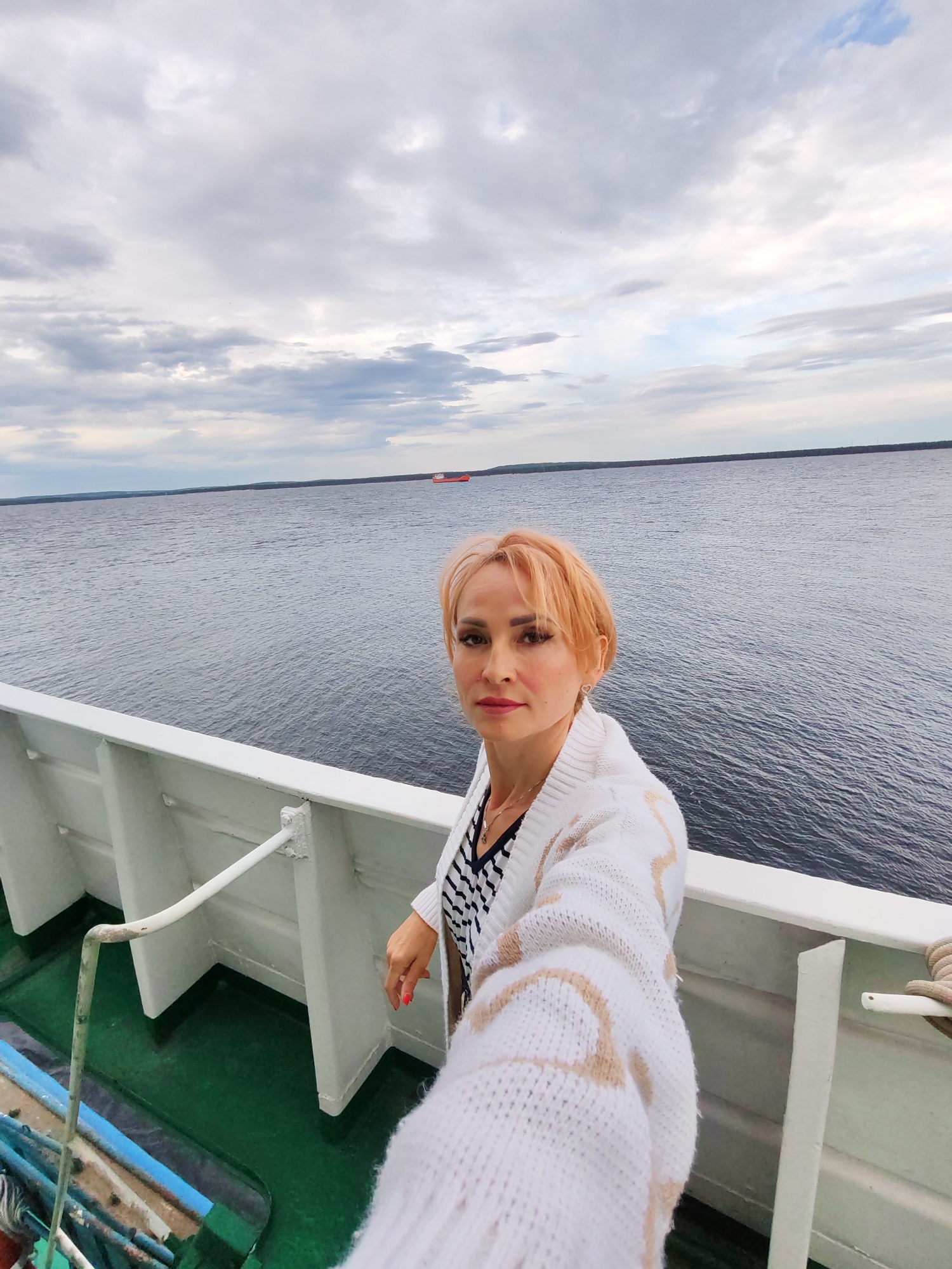 A woman with short blonde hair takes a selfie on the deck of a boat with the sea and a cloudy sky in the background. She is wearing a white cardigan with a tan pattern over a striped shirt. A red cargo ship is visible in the distance on the water. The deck of the boat is green, and there is a white railing in the foreground. The image appears to be taken during the day.