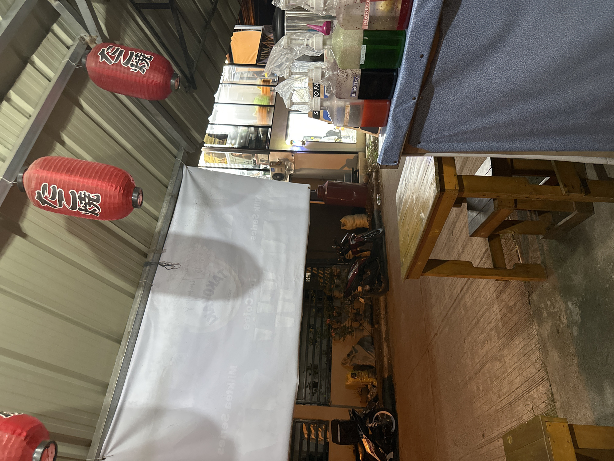 The image depicts an outdoor or semi-outdoor food stall or eatery, likely captured at night or during the late evening due to the dim lighting. The setting features a corrugated metal roof or awning.
Two red paper lanterns, bearing the Japanese characters "たこ焼" (Takoyaki), hang from the overhead structure. A long white banner or tarp stretches across the scene; reversed text visible on it includes "TAKOYAKI" and possibly "MILK SERIES" or "MILK COFFEE" when read in the correct orientation.
To the right, a counter holds several clear plastic bottles filled with colorful liquids, likely syrups or drink ingredients. Visible labels on these bottles denote "Four Seasons," "Blueberry," "Green Apple," and "Strawberry." Below the counter, a wooden table and benches are arranged for customers. The ground consists of rough concrete.
In the background, behind the white banner, parked motorcycles, some potted plants, and what appears to be an entrance to another building or part of the establishment are visible. The scene suggests a casual street-side or open-air food vendor, characteristic of urban environments like Davao City, Philippines. No individuals are present, so no specific activity is being performed, but the setup is indicative of food preparation and dining.