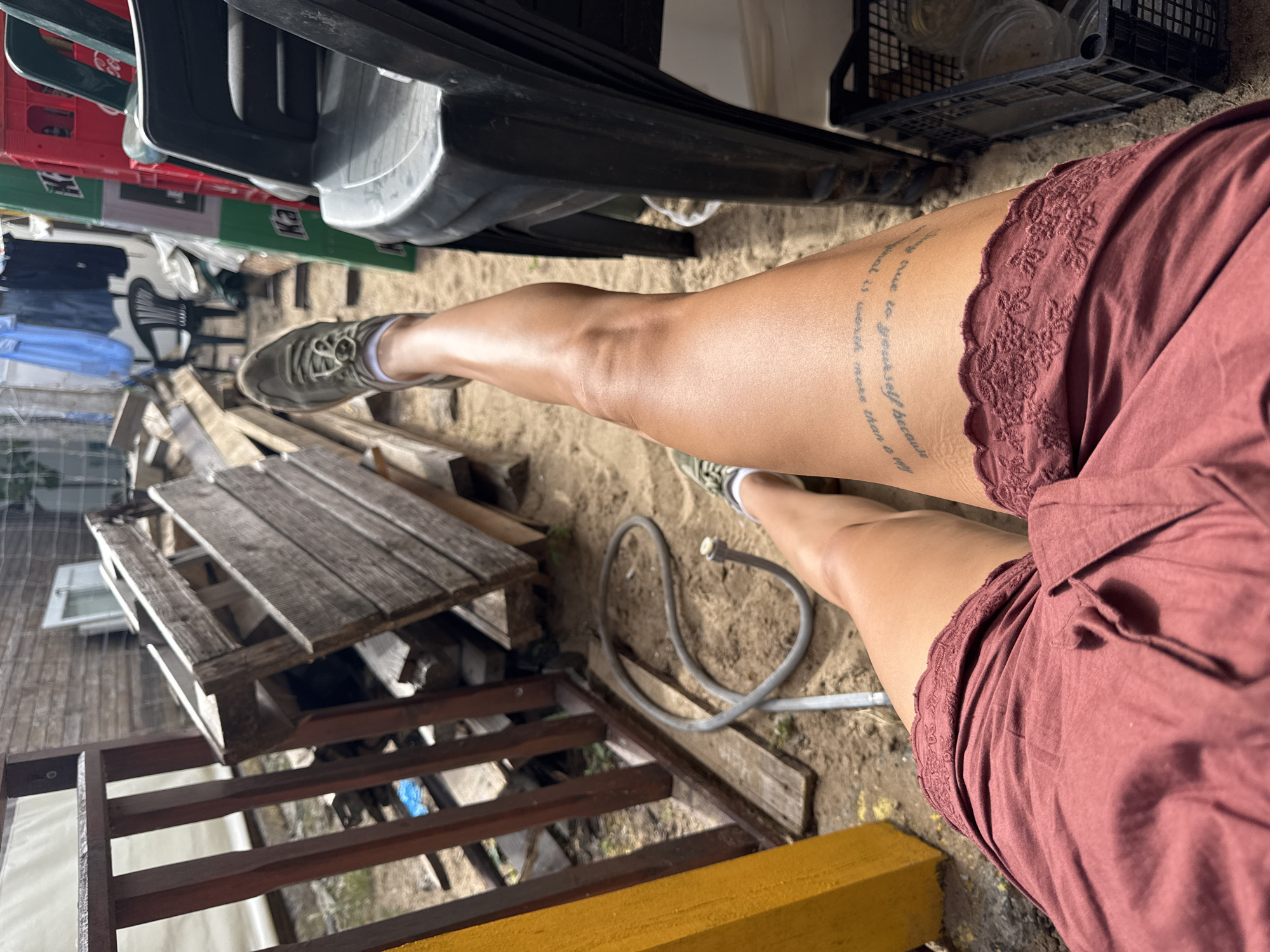 A person's legs are the primary focus of this media file, which appears to be taken outdoors. The individual is wearing a reddish-brown lace-trimmed skirt and green sneakers. A tattoo is visible on the person's left thigh, which encircles the leg and appears to contain text. The ground beneath the person is sandy, with some scattered wooden planks and a coiled hose visible. In the background, there are stacks of what look like plastic crates, some of which are red and green, and a wooden structure. The lighting suggests it is daytime. The scene gives the impression of a somewhat cluttered outdoor area, possibly a backyard or storage space. There are no clear indicators of a specific event taking place, but the image captures a moment in time. The location context of Sozopol, Bulgaria, is provided, but not directly evident from visual cues within the image itself.