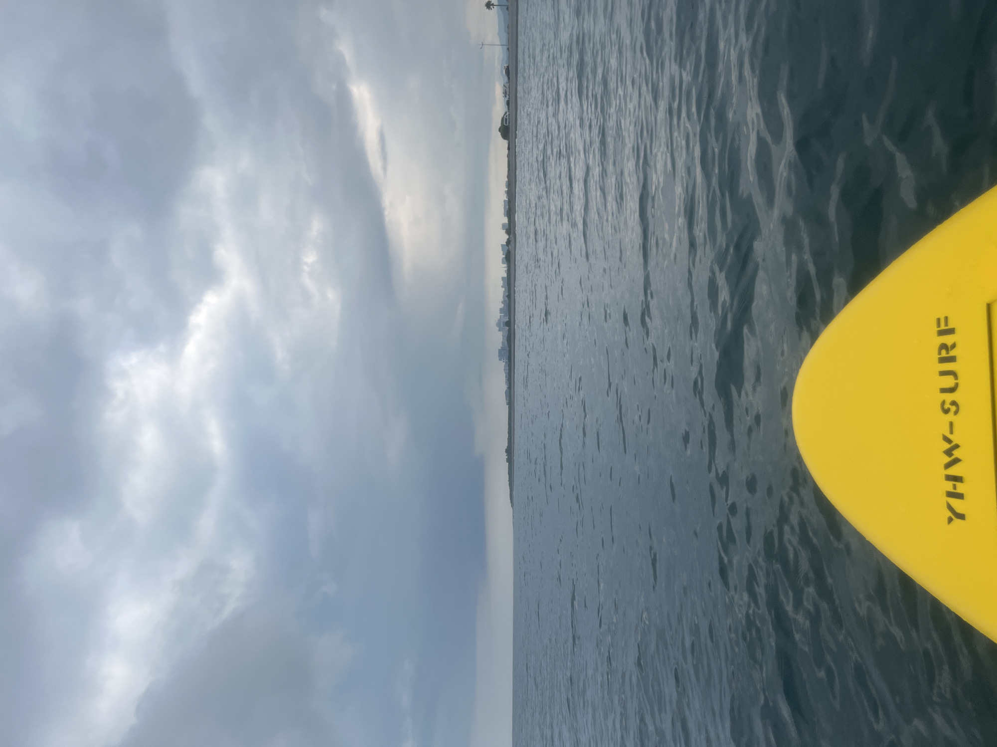 The image depicts a view from a yellow surfboard on the Black Sea, facing a coastline. In the distance, a city skyline, likely Batumi, Georgia, is visible along the shore. The sky is overcast with dramatic, dark clouds, suggesting the possibility of rain. The water is calm with gentle ripples. The surfboard has the text "YHW-SURF" printed on it. There are no people visible in the frame.