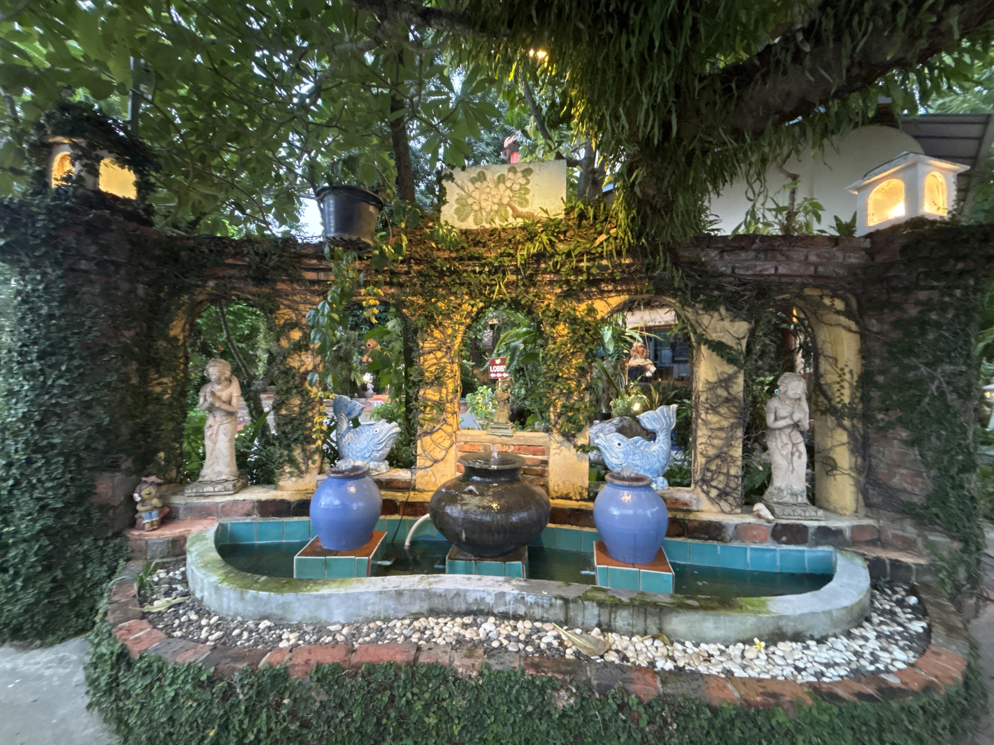 This is a medium shot of a tiered fountain and garden wall in Chalong, Thailand, in the evening. The wall is made of red brick and covered in green ivy. Three arched openings in the wall reveal glimpses of a garden beyond, lit with warm yellow lights. In the foreground, a circular fountain basin is filled with green water. Two blue ceramic pots with fish sculptures on top are placed on either side of a darker, larger ceramic pot in the center of the fountain. The fountain is surrounded by a decorative border of white pebbles and red brick. To the left and right of the fountain, there are two stone statues of women. A small, brown bear statue is visible to the far left of the fountain. The overall scene is lush and verdant, with the lighting creating a magical and inviting atmosphere.