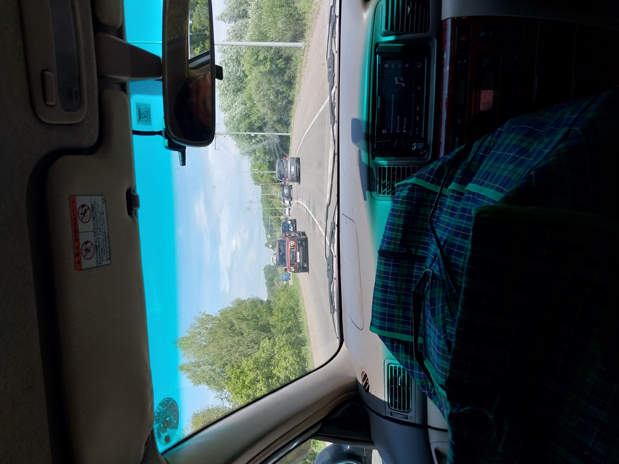 The image is a first-person view from inside a car, driving on a road with trees on either side. The sky is blue with some clouds. The dashboard of the car is visible, including the steering wheel and a radio display. A green and blue plaid shirt is draped over the passenger seat. In the distance, other cars are visible on the road. The scene appears to be set in a rural or suburban area. The text on the sun visor suggests a warning label.