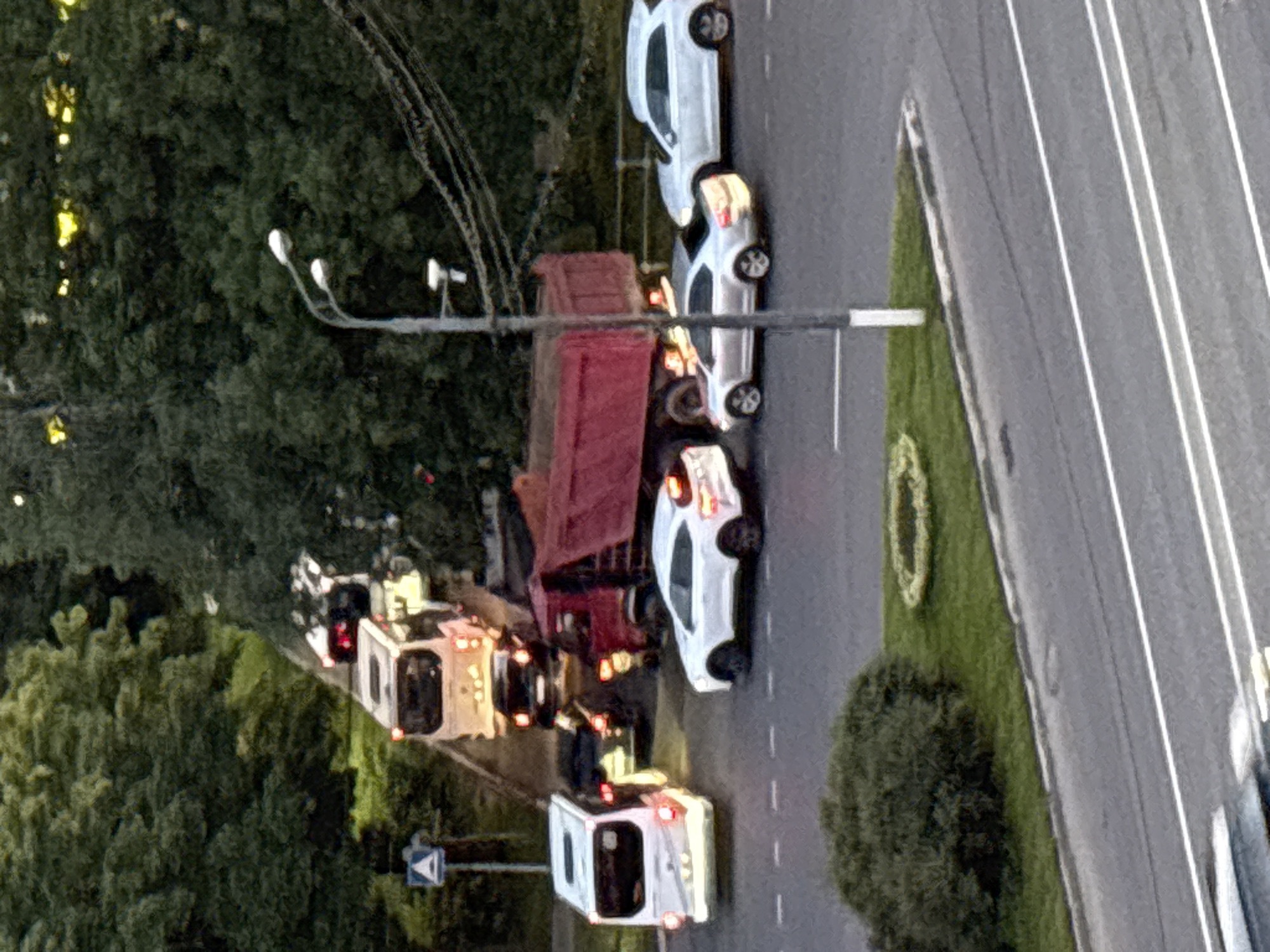 The image shows a traffic jam on a road during what appears to be the late afternoon or early evening, given the warm lighting and shadows. Several vehicles are visible, including a red dump truck, a white passenger car, and two white vans. The vans, one of which has its hazard lights on, suggest an emergency or breakdown situation. The scene is set against a backdrop of lush green trees, indicating an outdoor, possibly suburban or park-like setting. A road with clear white lane markings is prominent, suggesting a developed area. There are streetlights on a pole, further confirming an urban or semi-urban environment. No people are clearly discernible within the vehicles or the scene, but their presence is implied by the stopped traffic. The overall mood appears to be one of disruption and delay. There is no visible text in the image.