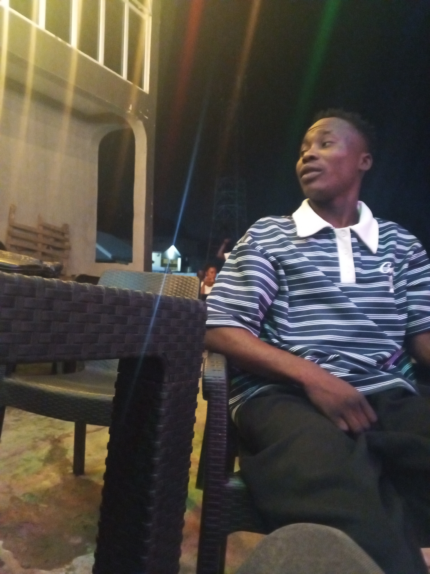 The image depicts a young man seated at an outdoor or semi-outdoor establishment in Lokoja, Nigeria, at night. He is positioned in the mid-ground, facing slightly to his right (viewer's left) with a neutral, observant expression. He wears a dark blue and white horizontal striped polo shirt with a white collar and a small, white embroidered logo resembling a stylized "C" on the left chest. His attire also includes dark trousers, and his right hand rests on his lap.
In the foreground on the left, a dark brown, woven rattan-style table is prominently featured, contributing to the depth of the scene. Similar rattan chairs are visible around the table and in the background. The setting appears to be a bar, restaurant, or lounge, indicated by the furniture and the relaxed ambiance. The background reveals parts of a building with modern architecture, including a white-framed balcony or upper level and a light-colored wall with an archway. Distant artificial lighting and distinct colored light beams (red, green, and blue) streak through the dark night sky, illuminating aspects of the environment and confirming the nighttime setting. A blurred wooden pallet or crate is also visible against a wall in the left background. Other indistinct figures are faintly discernible further in the background, suggesting a social gathering.