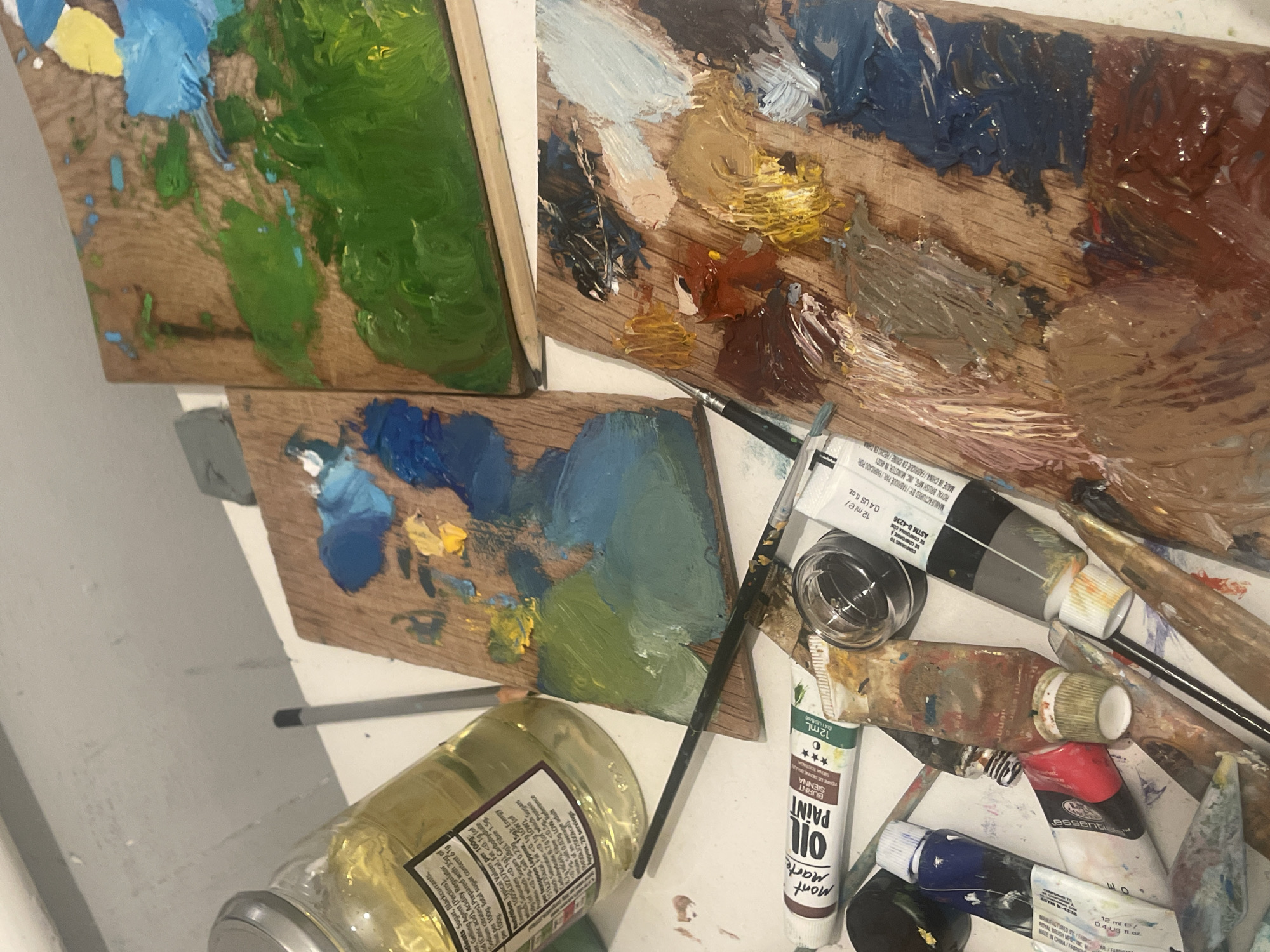 The image is a close-up shot of an artist's workspace, focusing on painting supplies. There are three wooden palettes covered in various colors of oil paint: blues, greens, browns, yellows, and whites. Several tubes of oil paint are scattered around, including a tube labeled "Mont Marte OIL PAINT Burnt Sienna." Paintbrushes and a small container (possibly for cleaning brushes) are also present. A jar filled with a yellow liquid is in the lower left corner; the label on the jar contains nutritional information, suggesting it may be a food item repurposed for art use. A white surface serves as the backdrop for the scene. The information extracted from the OCR text indicates that the art supplies are manufactured by Royal Brush MFG, INC. in Munster, IN 46321 and conform to ASTM D-4236 standards. There is no mention of Maidstone, United Kingdom, or any people or actions in the image.