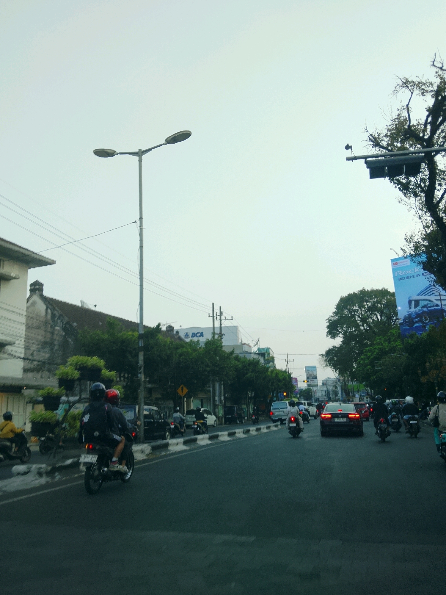 The image captures a street scene in Malang, Indonesia, likely during the day given the ambient light. A prominent double-headed streetlamp stands on the left side of the road, its poles extending upwards against a pale sky. Across the street, buildings with varying architectural styles line the road. On the right side, large trees, some bare of leaves, frame the scene. A billboard advertising "Rock" and "BELIEVE IN R" is visible further down the road on the right.
The street itself is a busy thoroughfare with traffic. Several motorcycles are present, with two individuals on one motorcycle in the foreground, wearing helmets and backpacks. Other motorcycles and cars are dispersed throughout the road, indicating moderate traffic flow. A white van is parked on the left side. Traffic lights are visible in the distance. The road surface is asphalt, with painted lane markings. The overall atmosphere suggests an urban environment with ongoing daily activities.