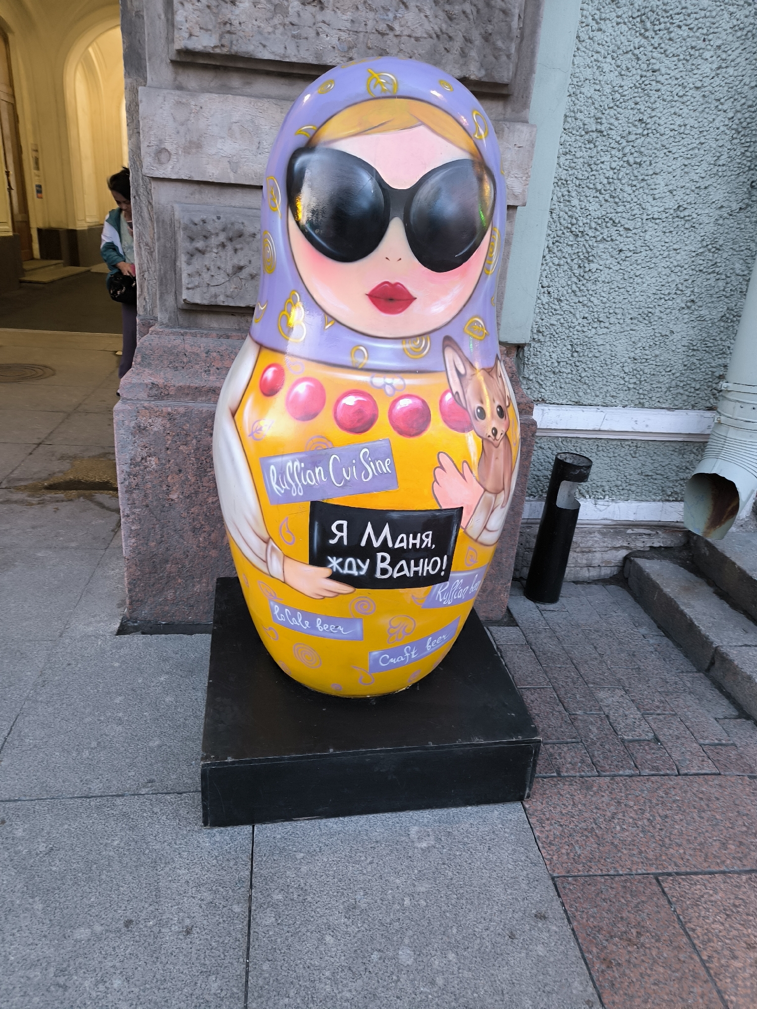 A large, vibrantly painted Matryoshka doll is displayed outdoors on a black pedestal in Saint Petersburg, Russia. The doll is decorated with a purple headscarf, oversized black sunglasses, and rosy cheeks. It holds a small sign that reads "Я Маня, жду Ваню!" (I am Manya, waiting for Vanya!) in Russian. Various other inscriptions, such as "Russian Cuis Sine," "Caft beer," and some floral patterns, adorn the doll's yellow body. To the right of the doll, a black cylindrical object, possibly an ashtray or trash receptacle, stands on the pavement. Behind the doll, a textured, light-colored wall rises, and to the left, a stone archway with a person partially visible entering it can be seen. The ground is paved with granite tiles.