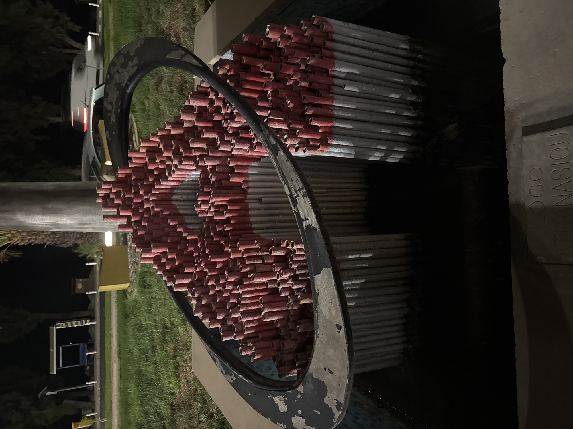 In the city of Germasogeia Municipality, Cyprus, an outdoor sculpture is depicted at night. The sculpture is composed of numerous cylindrical elements, predominantly silver with red tips, arranged in a dense, tiered formation. A black, oval metal frame encircles the upper portion of the sculpture. The base of the sculpture appears to be a shallow pool of dark water, with some of the cylindrical elements submerged. In front of the sculpture, a stone plaque is visible with engraved text that appears to read "LOGO" and "ATHANASIOU". The background shows a dimly lit grassy area with trees and a palm tree. Further in the background, across a street, there are some structures including a bus stop and a parked car. The overall lighting suggests it is nighttime, with artificial lighting illuminating the sculpture and its immediate surroundings.