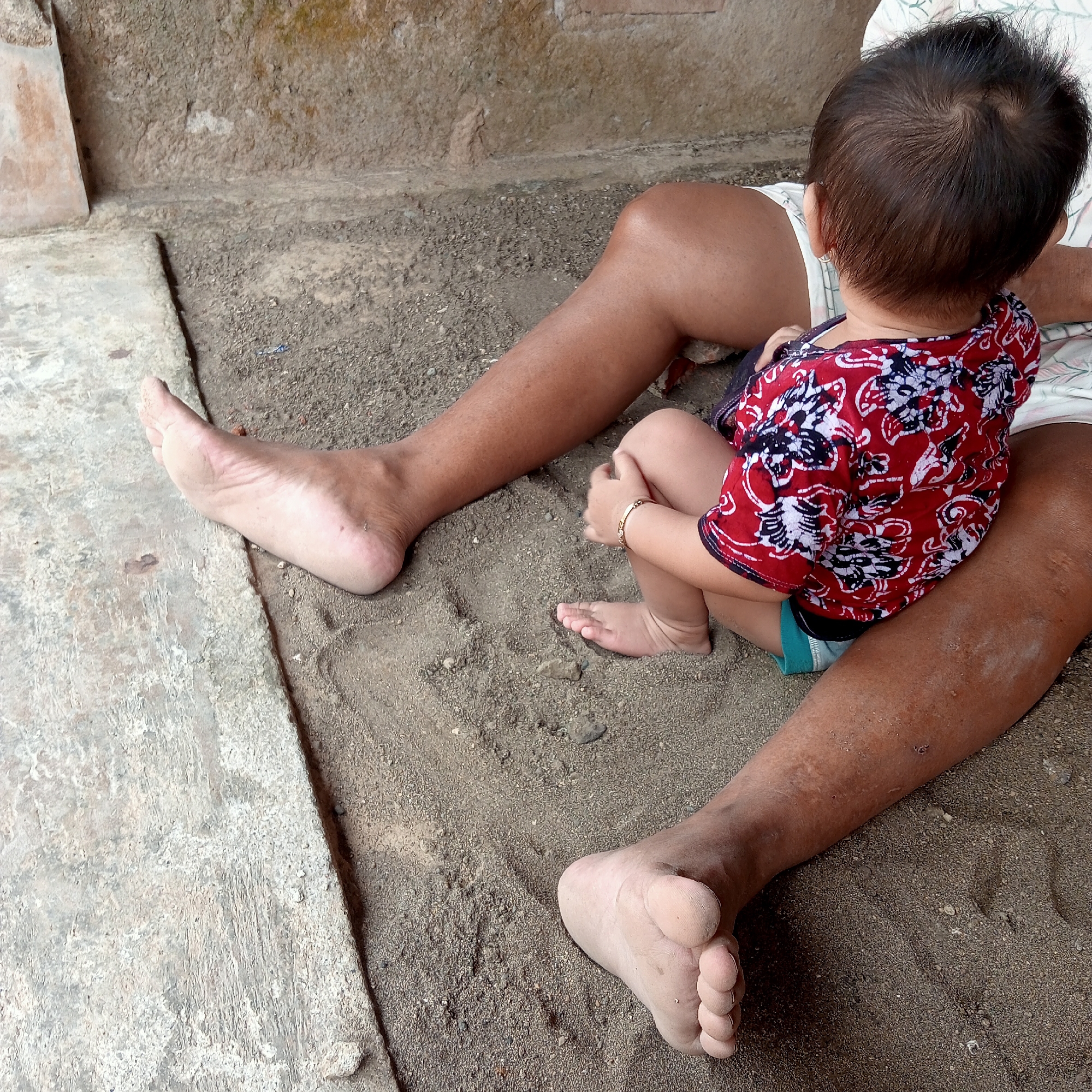 A baby is sitting on the lap of an adult, who is lying down on a sandy ground. The adult's legs are spread apart, and the baby is facing away from the camera. The baby is wearing a red and white patterned shirt and a gold bracelet on its wrist. The adult is wearing white shorts. The environment appears to be outdoors, possibly in a rural or undeveloped area, given the sand and the rough-textured wall in the background. The lighting suggests it is daytime. The overall scene depicts a moment of rest or play between an adult and a child. The location is specified as Bangsri, Indonesia.