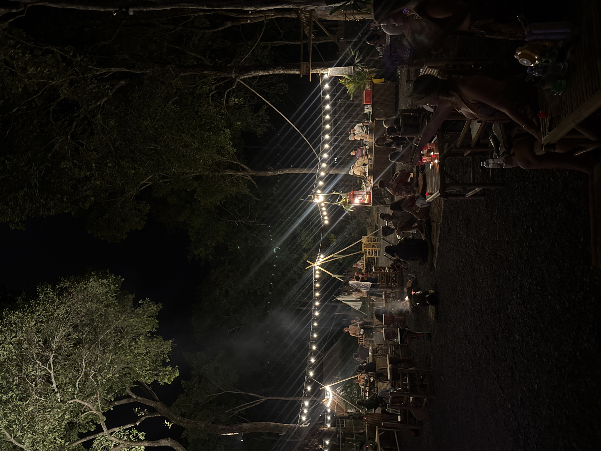 The image captures a nighttime outdoor gathering in ตำบลเกาะพะงัน, Thailand, set within a wooded area. Numerous tall trees with dark foliage frame the scene and rise into a black sky. The ground is dark, appearing to be gravel or packed earth.
The central area is illuminated by multiple strings of bare bulb lights suspended across the space, casting visible light rays. A small campfire burns in the mid-ground, surrounded by several Djembe drums. Approximately 30 to 40 individuals are visible, engaged in various activities.
People are seated at rustic bamboo tables and chairs, or directly on mats on the ground, socializing. On the right side, a long bamboo counter serves as a food or drink station, with several individuals, including staff, working behind it. A red popcorn machine is visible on this counter. In the foreground on the right, a woman with light-colored hair is seated at a bamboo table, extending her right arm forward. She wears a patterned top and shorts. Her table holds a plastic water bottle, small lit candles, and other items. Another woman is partially visible next to her.
In the mid-ground, a shirtless man stands centrally, observing the activities. Other individuals are gathered around the campfire and near the bamboo structures, some appearing to be engaged with the drums. Various bamboo-pole structures, including teepee-like frames, are interspersed among the seating areas. The overall atmosphere is that of a casual, communal outdoor event.