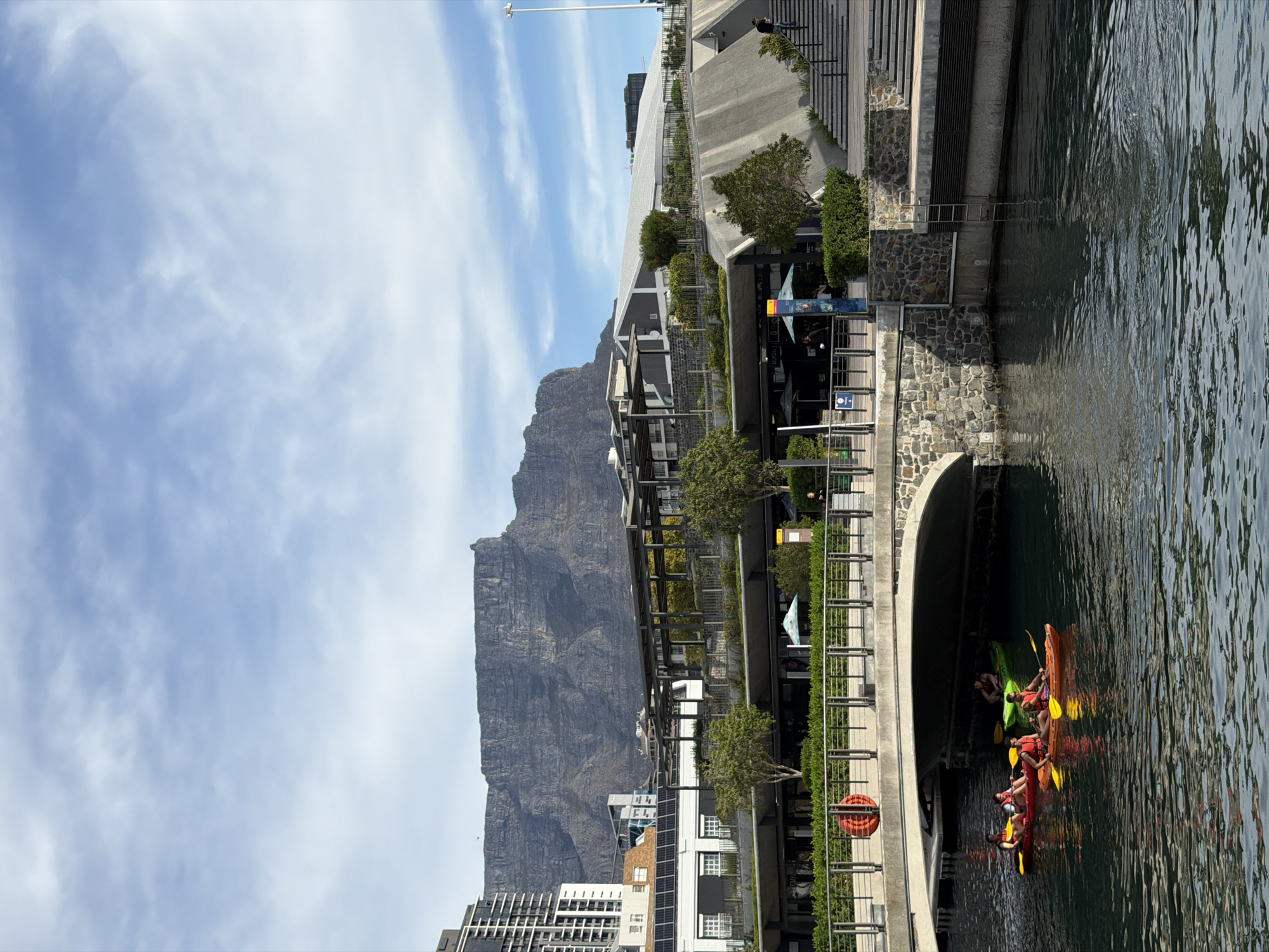 The image shows a waterfront scene in Cape Town, South Africa, with Table Mountain in the background. The V&A Waterfront is visible in the foreground, with modern buildings, landscaping, and a canal. People are kayaking in the canal. The sky is blue with scattered clouds.