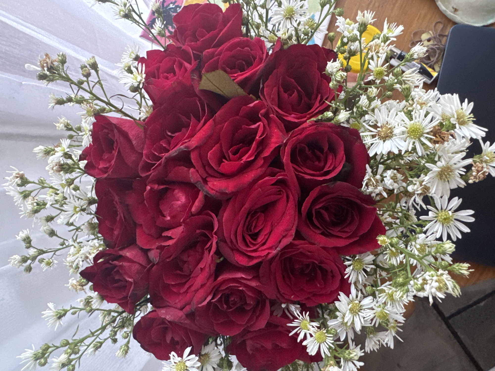 A bouquet of deep red roses and delicate white filler flowers is arranged on a wooden surface. The bouquet is viewed from above, with the roses dominating the center of the frame. The white flowers, characterized by their small, daisy-like petals and green stems, surround and cascade around the roses. Some of the white flowers appear slightly wilted. The background is a soft, white, draped fabric, possibly a curtain or tablecloth. The wooden surface appears to be a table, with glimpses of dark tiles or flooring visible at the bottom left. A dark electronic device, possibly a laptop or tablet, is partially visible in the bottom right corner of the frame. There are no people or animals present.