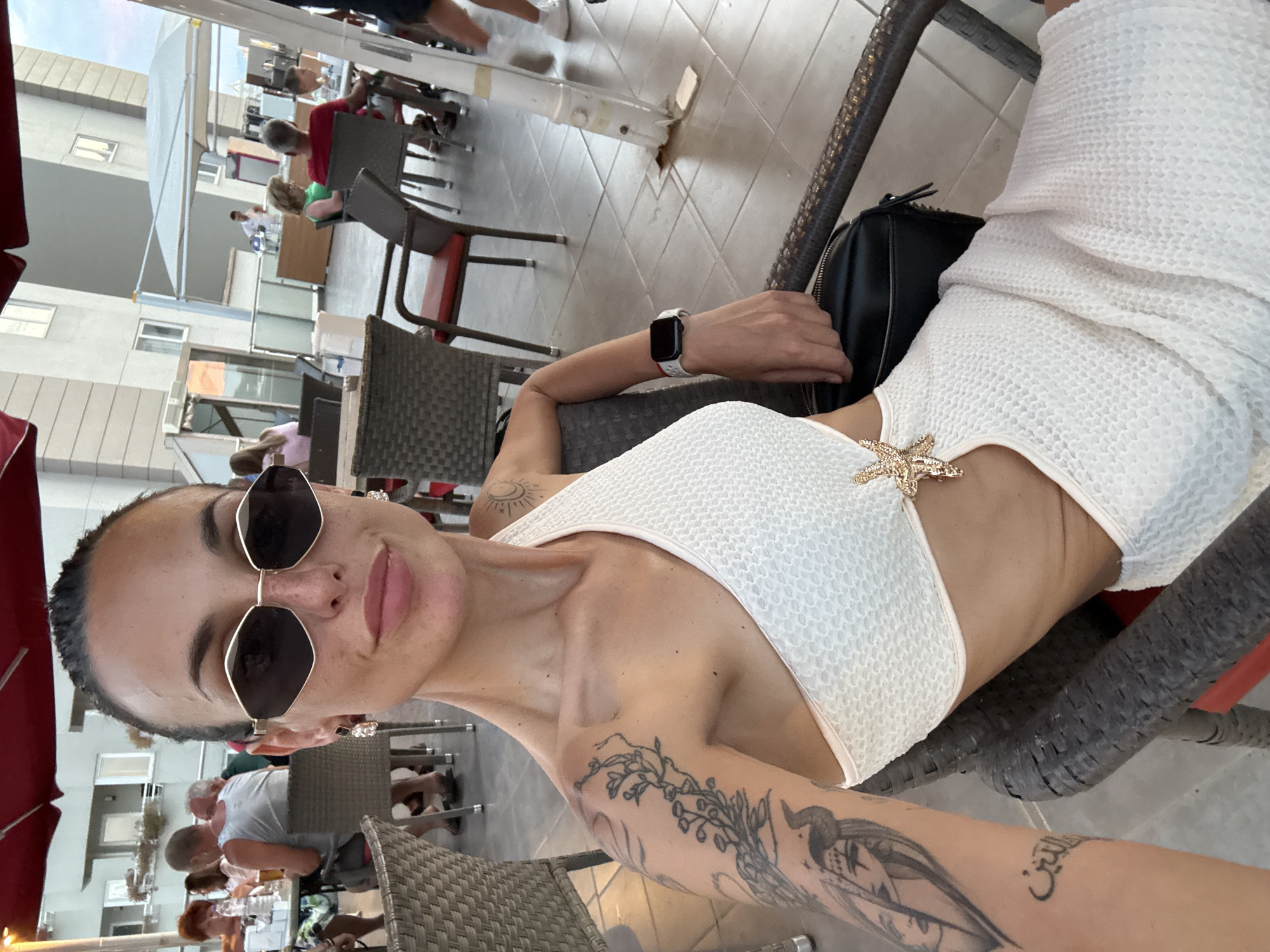 A woman with dark hair, pulled back, and sunglasses is taking a selfie in an outdoor seating area, likely a cafe or restaurant. She is wearing a white, one-shoulder dress with cutouts. A gold starfish ornament is attached to the dress. Tattoos are visible on her left arm and shoulder. She is wearing a watch on her left wrist. In the background, other patrons are seated at tables, and a building with balconies is visible. The floor is tiled. This scene is located in Aksu, Turkey.