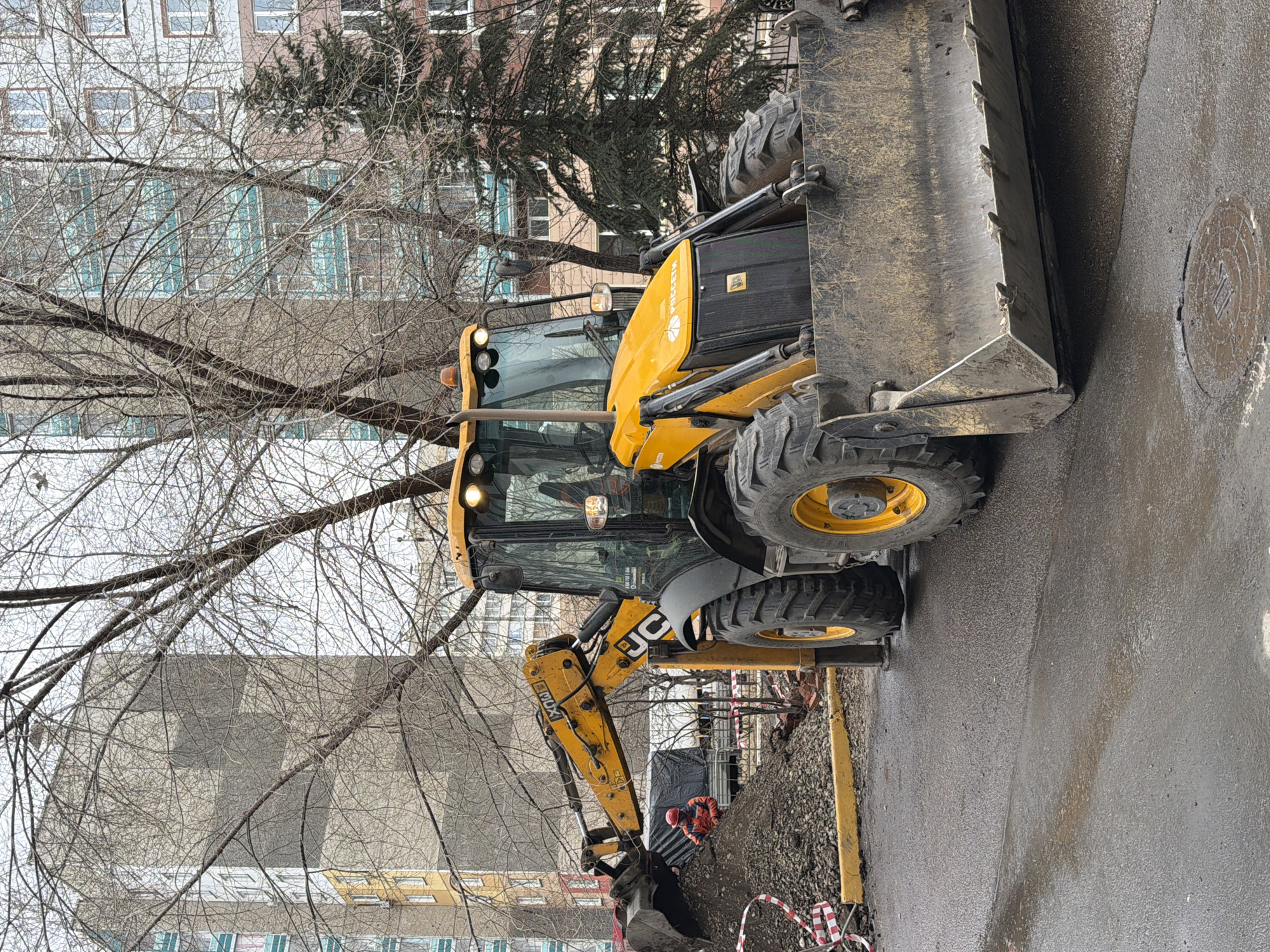 A yellow JCB backhoe loader is parked on a wet asphalt road in a residential area. The backhoe's front bucket is raised. A person wearing an orange jacket is working near the backhoe's rear bucket on a pile of dirt marked off with red and white striped tape. Bare trees are in the background, along with apartment buildings. A manhole cover is visible in the upper right corner of the image. The location is likely in Krasnoyarsk, Russia.