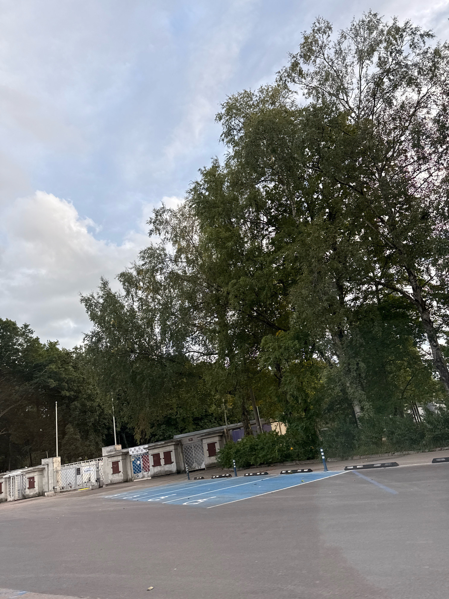 The image depicts an outdoor scene featuring a grey asphalt parking lot with designated parking spaces marked in blue. To the left of the parking lot, there is a low, light-colored concrete structure with a metal fence in front of it, behind which are several small buildings with red doors and windows. Beyond the structures, a dense line of green trees and foliage forms a backdrop. The sky above is overcast with scattered clouds. In the foreground of the parking lot, there are black parking stops. The overall scene is devoid of people. The image appears to be taken in Tallinn, Estonia, suggesting it could be a public parking area, possibly near a sports facility or park given the presence of the fence and buildings.