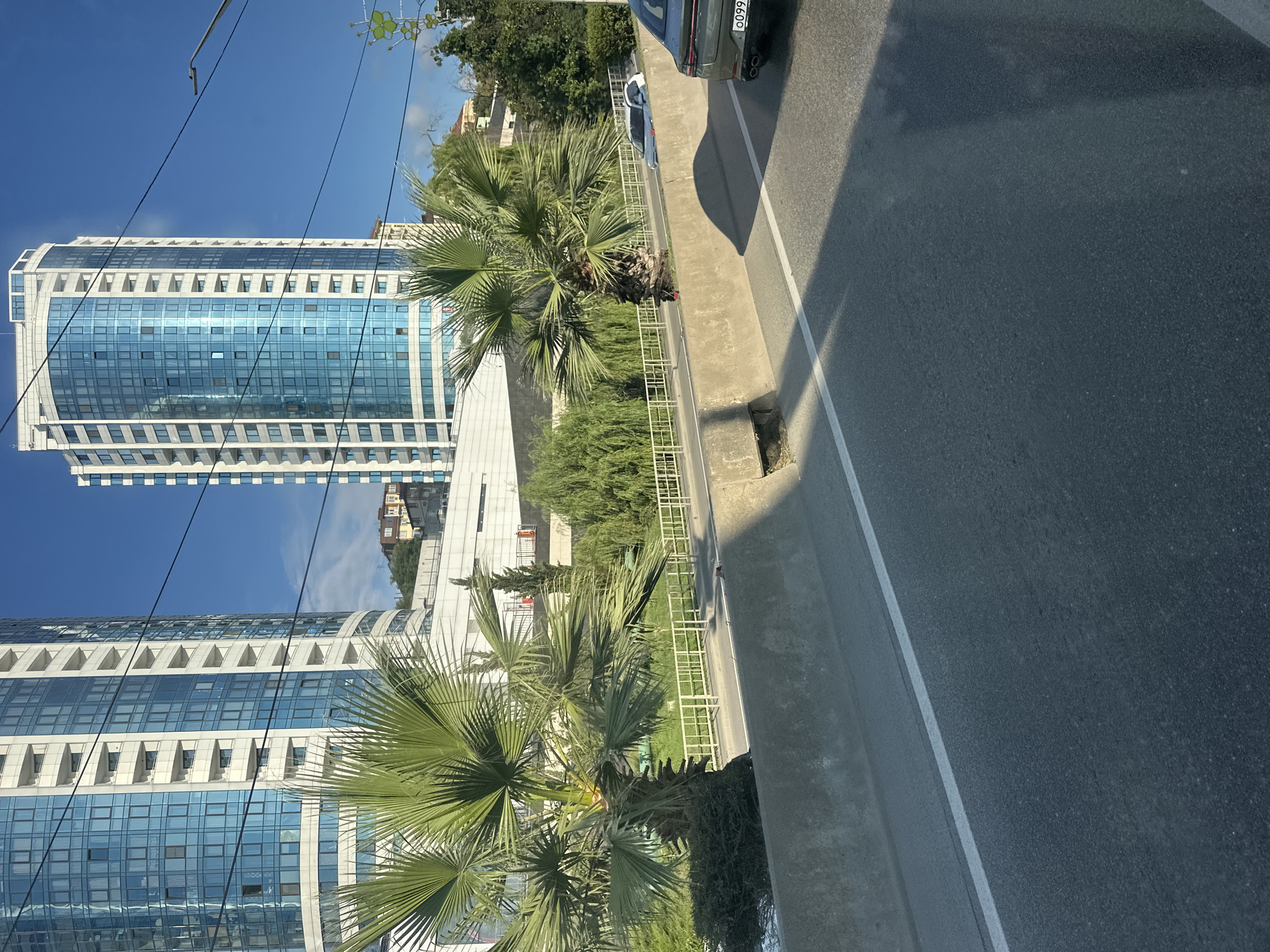 The image depicts a street scene in Sochi, Russia, with modern high-rise buildings in the background and a roadway in the foreground. Two prominent skyscrapers with blue glass facades and white balconies dominate the upper portion of the frame. In front of the buildings, there are palm trees and lush greenery. The roadway has a concrete barrier on the right side, with a white line marking the lane. Two cars are visible on the roadway, one light-colored and partially obscured, and another dark-colored car with its license plate visible. The overall environment suggests an urban setting with elements of a warm climate, given the presence of palm trees. The sky is clear blue.