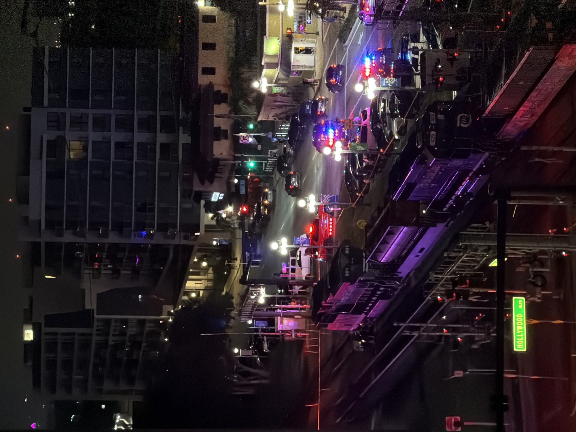 At night in Hollywood, Florida, a train passes by a scene with multiple law enforcement vehicles, including police cars with flashing lights. The train, a dark locomotive with purple lighting along its side, is on tracks in the foreground. In the background, a street is visible with numerous cars, some parked and others in traffic. Buildings line the street, including apartment complexes with illuminated windows and a lower-rise building with shops. The overall scene suggests an active law enforcement presence on a city street at night. A street sign reads "HOLLYWOOD BLVD."