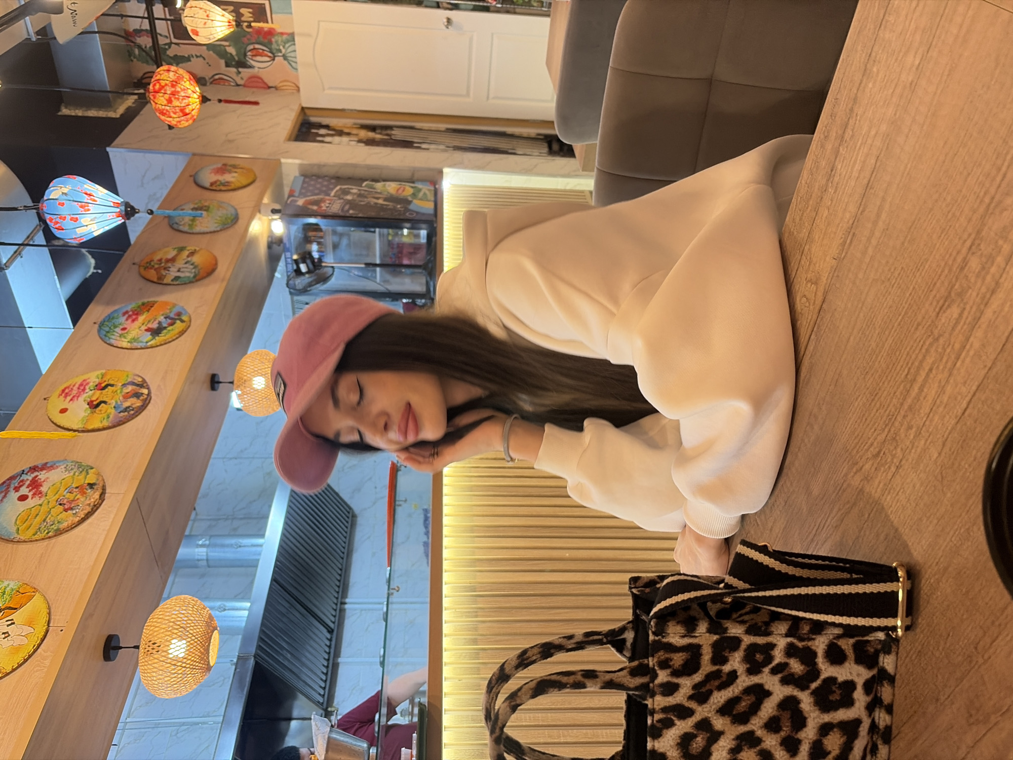 A young woman, wearing a pink baseball cap and a white hooded sweatshirt, is captured in a moment of relaxation, resting her head on her hand at a wooden table. Her eyes are closed or partially closed, suggesting a calm or contemplative state. A distinctive leopard print tote bag with black and white striped handles is placed on the table beside her.
The setting appears to be an indoor restaurant or cafe, likely in Izhevsk, Russia, with an East Asian-inspired decor. The space is illuminated by warm, ambient light emanating from various decorative lanterns of different shapes and colors, suspended from the ceiling. Along a wooden beam or shelf on the wall, several circular decorative plates featuring colorful, intricate designs are displayed.
In the background, upholstered seating, possibly booths or chairs, can be seen, along with a white paneled door. Further into the background, elements of a kitchen area, including a metallic ventilation hood, are visible. The overall atmosphere is warm and inviting. No readable text is discernible within the image.