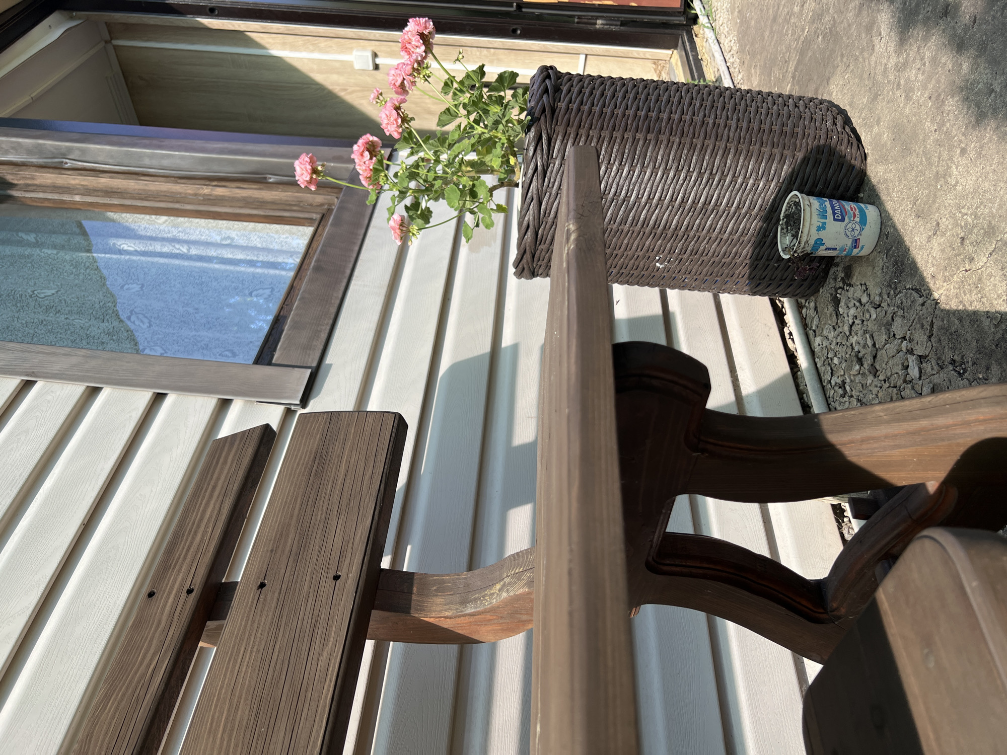 In the city of Tashelka, Russia, a wooden bench sits beside a building with white horizontal siding. In front of the building, a large, dark brown woven planter holds a flowering plant with pink blossoms. To the right of the planter, a small, white plastic cup with blue lettering is on the ground. The scene is illuminated by sunlight, casting shadows.