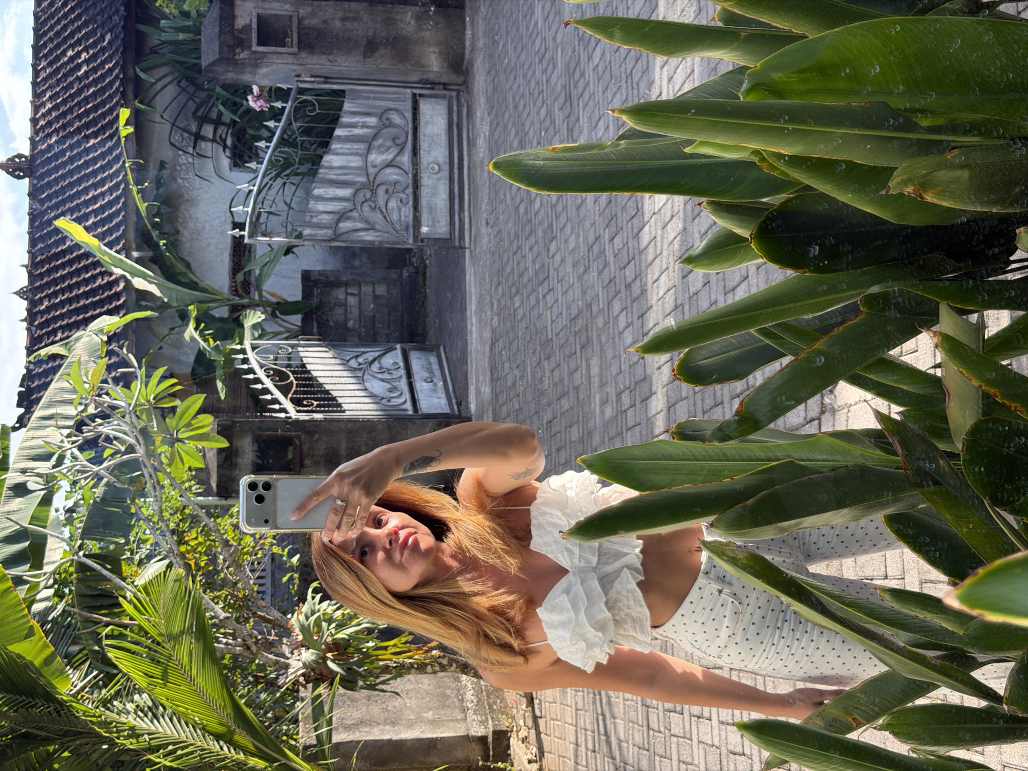 A woman with shoulder-length auburn hair is taking a selfie in a paved courtyard. She is wearing a white ruffled crop top and white pants with a dark polka dot pattern. Her right hand is raised, holding a silver smartphone to take the photo. A tattoo of a crescent moon is visible on her left forearm. She is standing in front of lush green foliage. In the background, there is a house with a dark tiled roof and a decorative metal gate. The setting appears to be outdoors, possibly in a residential area of Kuta Selatan, Indonesia, given the architectural style of the house and gate, as well as the tropical vegetation. The ground is paved with rectangular grey bricks. Sunlight is casting shadows, indicating it is daytime.