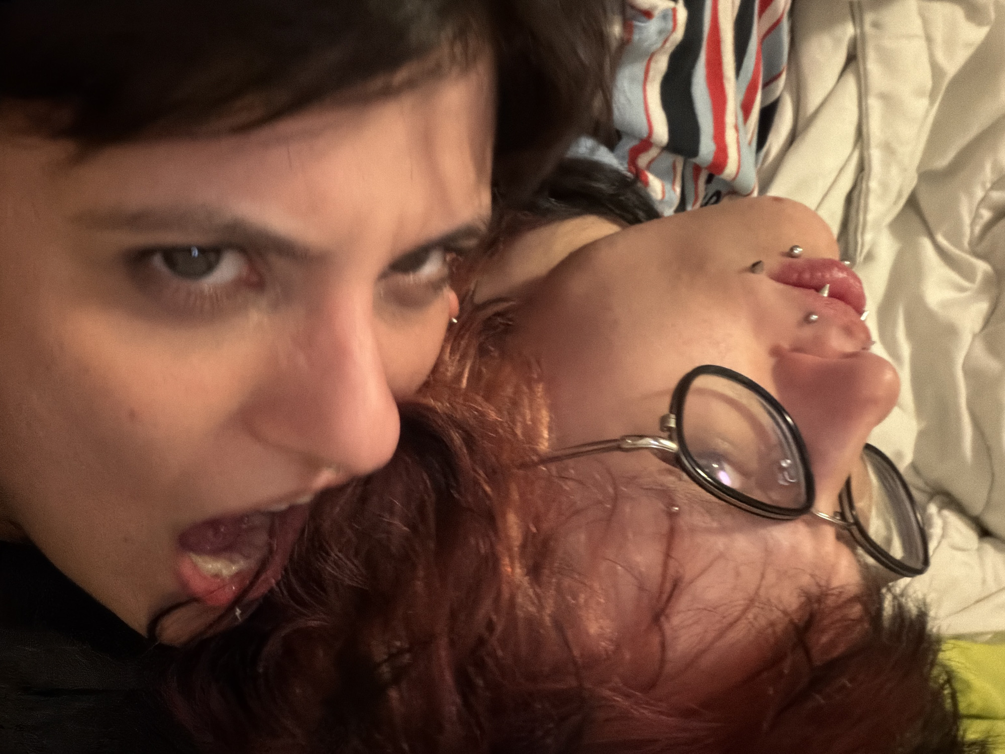 Two individuals are in close proximity. One individual with dark hair and eyes has their mouth open and appears to be biting or approaching the other individual's head. The second individual has red hair and is wearing round glasses. They have multiple piercings on their tongue. The background includes a striped garment and a white quilted surface. The location is not determinable from the visual information.
