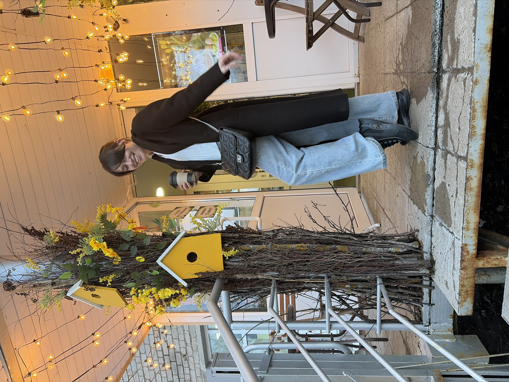 The image shows a person in front of a storefront. The individual is wearing a black coat, light blue jeans, and black shoes, and is holding a coffee cup and a black quilted bag. The storefront is decorated with string lights and a display of yellow birdhouses attached to branches and flowers. The location appears to be in Chaikovsky, Russia. The person is smiling and gesturing with their arm. The ground is paved and there are stairs and metal railings visible.