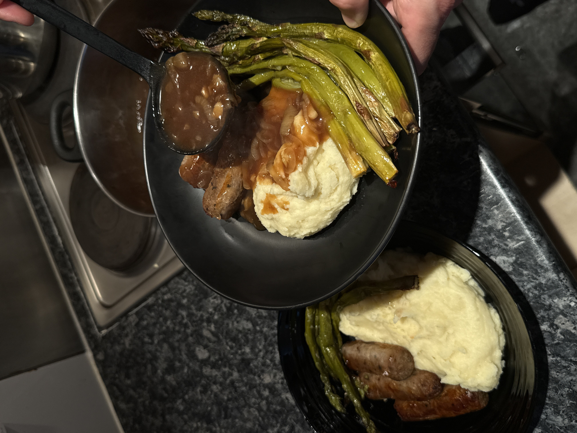 The image shows a plate of sausage and mash with asparagus being topped with gravy. A black ladle is pouring gravy onto a black bowl containing cooked sausages, a mound of mashed potatoes, and several spears of cooked asparagus. Another black bowl containing similar ingredients sits on a dark, speckled countertop. A stainless steel sink and cooking pot are visible in the background. The location cannot be identified from this image.