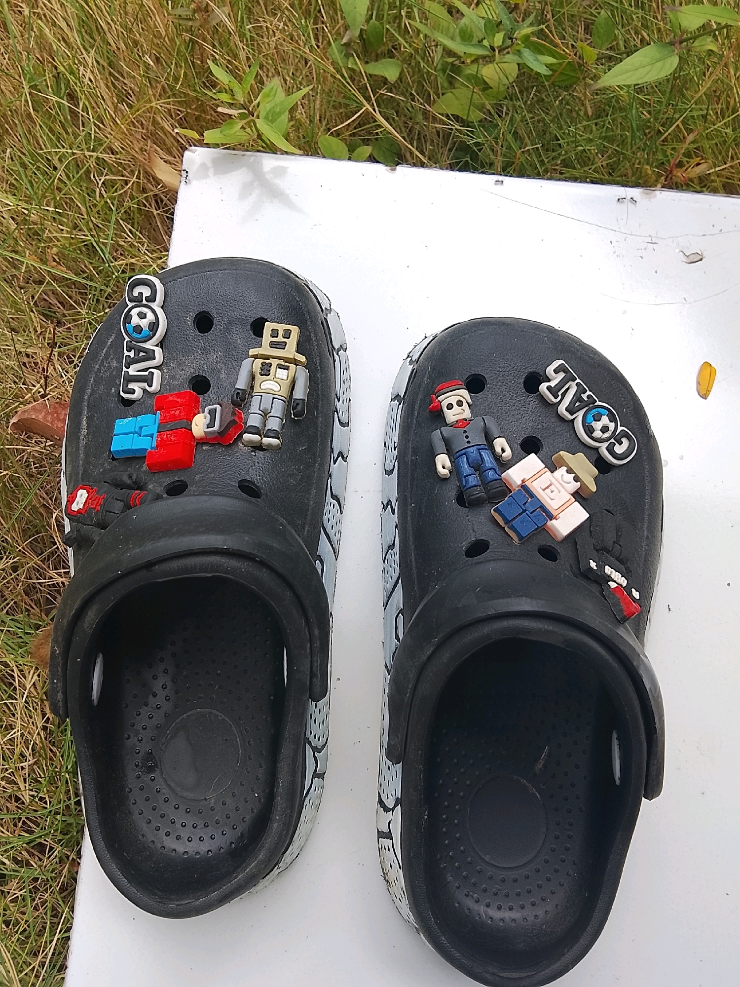 Two black Crocs, decorated with various jibbitz charms, rest on a white surface outdoors. The Crocs feature charms depicting a soccer ball with the word "GOAL", robots, and figures resembling Roblox characters. The left shoe has charms of a robot, a stack of blue and red blocks, and a red and black object. The right shoe has charms of a figure in a red hat, a figure with a tan hat, and a red and black object. The background consists of dry grass and green foliage, suggesting an outdoor setting. There is no indication of Kertahayu, Indonesia.