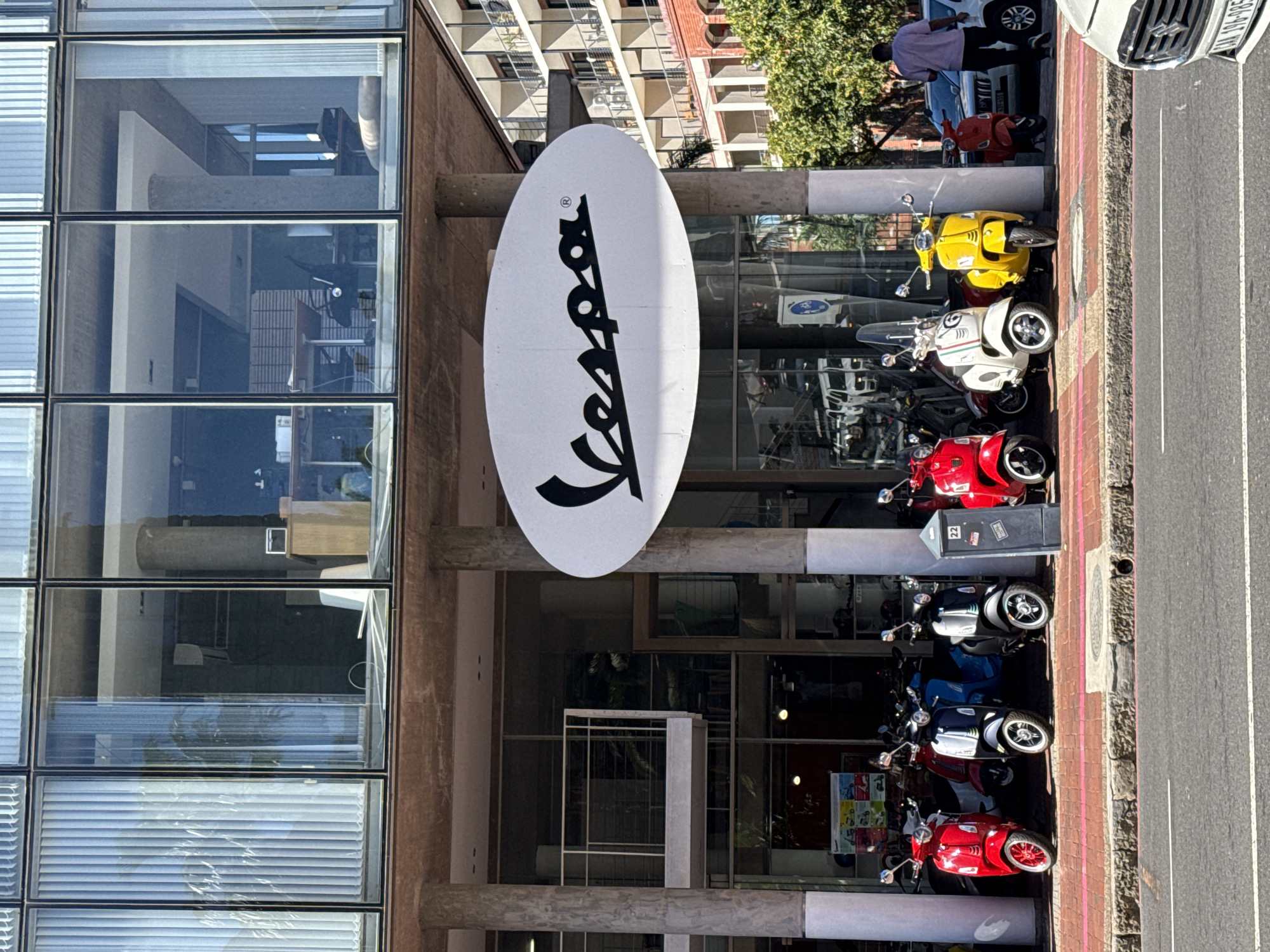 The image displays a street-level view of a modern building housing a Vespa scooter dealership in Cape Town, South Africa. A large, oval white sign bearing the black "Vespa" logo is prominently mounted above the ground-floor showroom.
On the red-brick sidewalk beneath the building's overhang, numerous new Vespa scooters are parked in a line, exhibiting various colors including red, dark grey, black, white with green and red stripes, and yellow. Additional scooters are visible through the large glass windows of the showroom. A dark grey utility box, marked with the number "22," is positioned on the sidewalk among the parked scooters.
In the background to the right, a multi-story residential building with multiple balconies and a brick base is partially visible, interspersed with green tree foliage. The upper floors of the dealership building consist of offices or apartments with large glass windows, some featuring vertical blinds.
On the asphalt street in the foreground, a white Suzuki vehicle is partially visible on the right, displaying a South African license plate "CA 196-985," confirming the Cape Town location. Further right, an adult male, seen from behind, walks leftward on the sidewalk, wearing a light shirt and dark trousers. Another white vehicle is parked behind the man.
