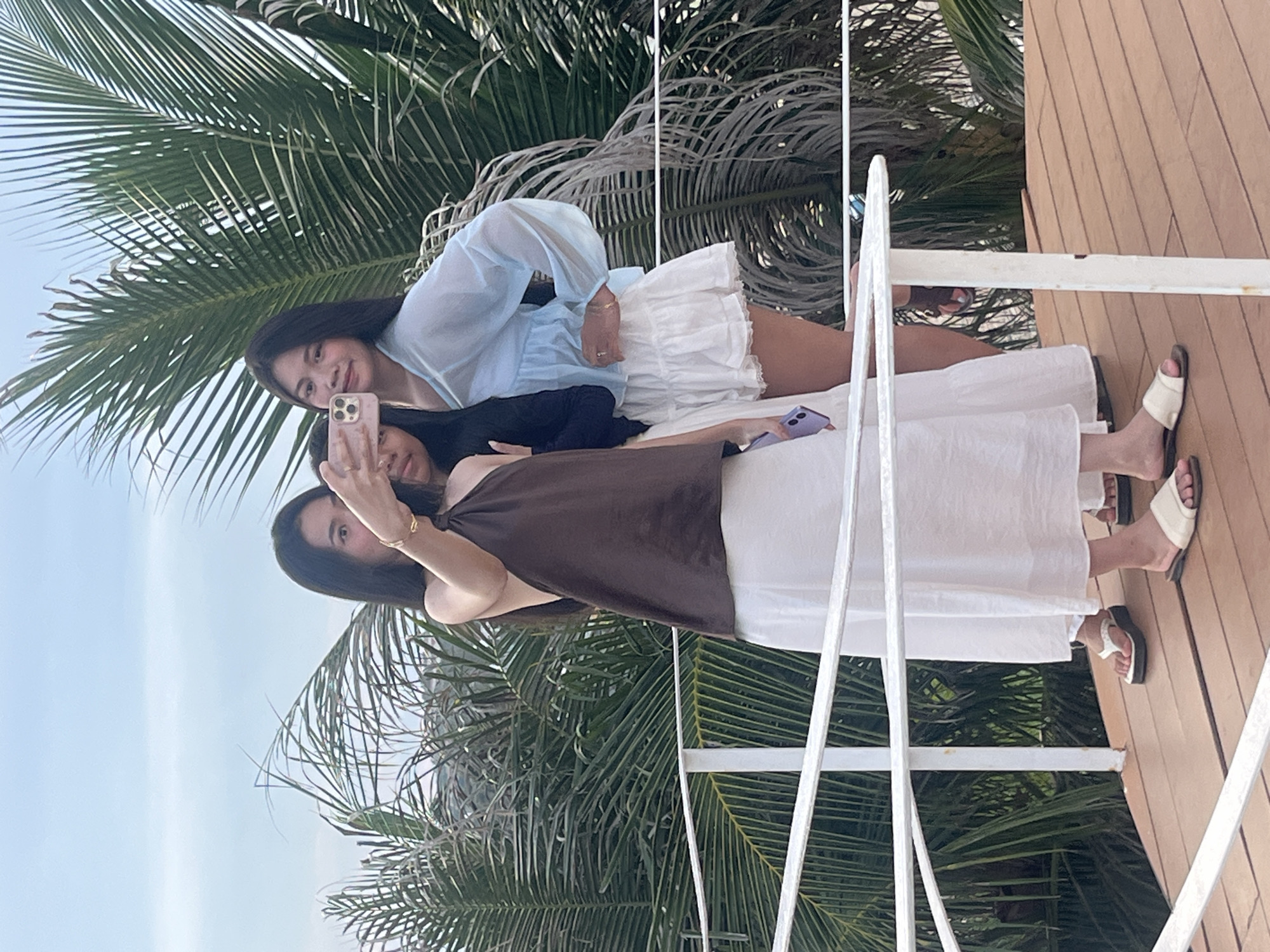 Three young Asian women are taking a selfie on a wooden platform with a white railing, in Mũi Né, Vietnam. The woman on the left is wearing a brown halter top and a long white skirt. She is holding up a phone to take the picture. The woman in the middle is wearing a black top and is partially obscured. The woman on the right is wearing a light blue sheer blouse and a short white ruffled skirt. She has her arm around the woman in the middle and is smiling. They are standing in front of lush green palm trees, with a cloudy sky in the background. The scene appears to be a scenic overlook or a resort area in Mũi Né.