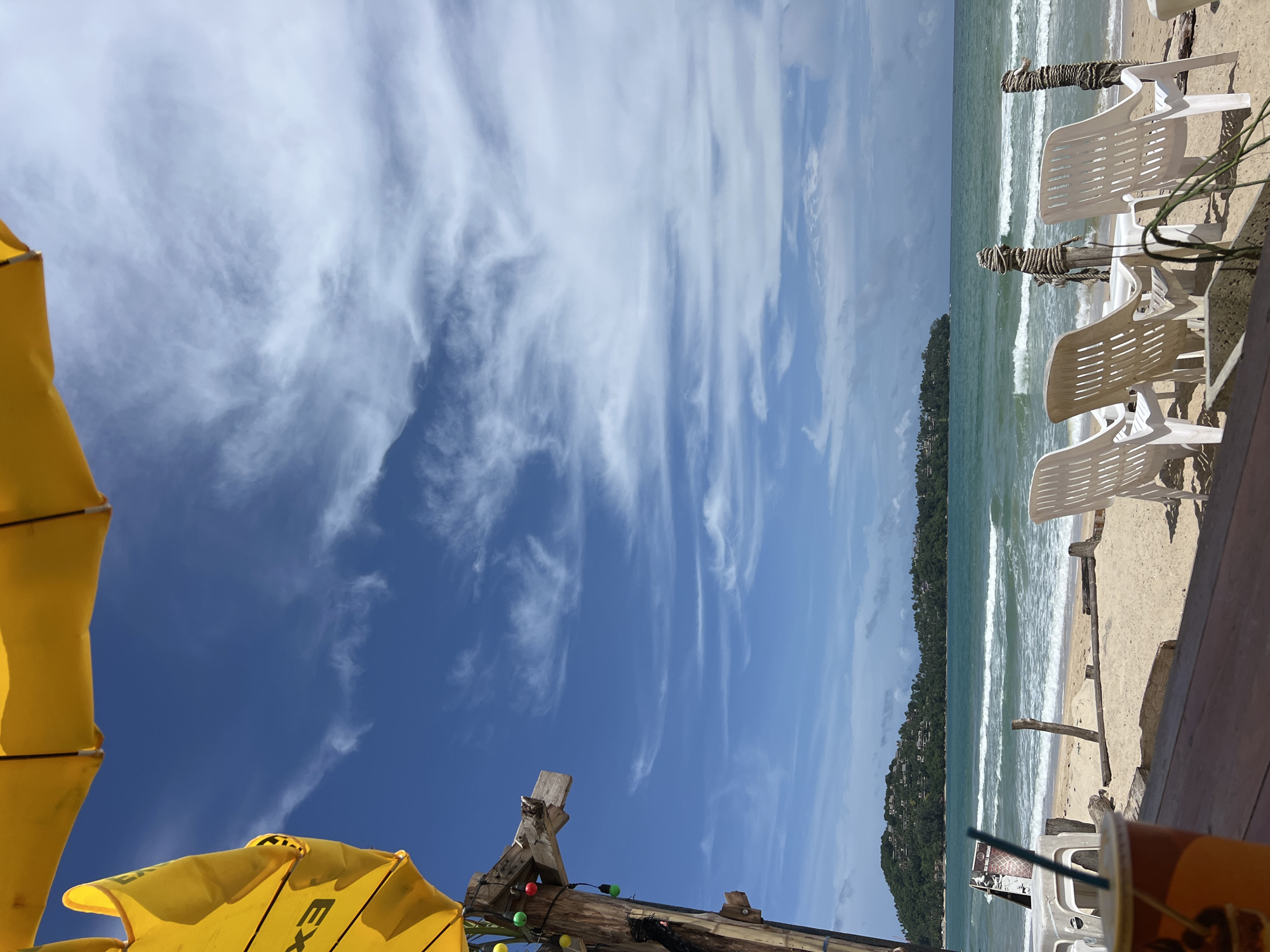 An outdoor perspective captures a beach scene under a bright, partly cloudy sky. In the foreground, a wooden table surface is visible, with a partially obscured orange cup and a straw on its left side. Further back, arranged on the sand, are several white plastic beach chairs, some facing the ocean. Two wooden posts wrapped in rope stand in the sand between the chairs and the water. The ocean exhibits moderate waves and a greenish-blue hue, meeting a sandy beach dotted with driftwood. In the distance, a tree-covered island with scattered buildings rises from the sea, and the horizon line is clear. Above, a large yellow umbrella stretches across the top of the frame, casting a shadow. A wooden pole with decorative lights is positioned to the left of the frame, attached to what appears to be a structure providing shade. The overall atmosphere suggests a relaxed, tropical setting.