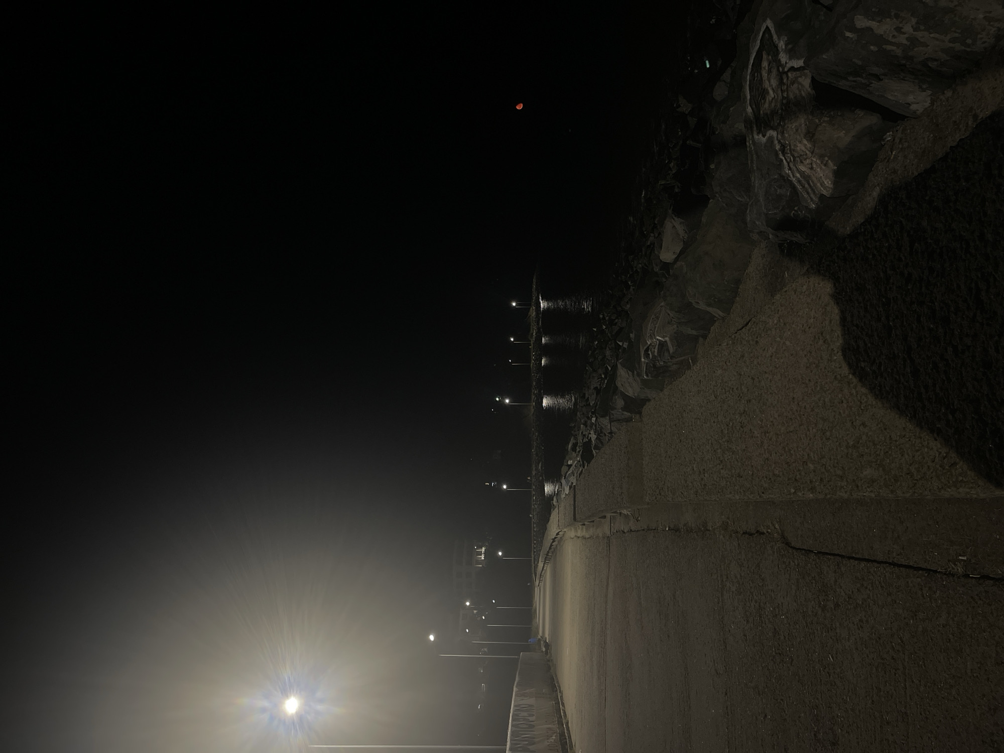 The image depicts a dimly lit walkway along a rocky shoreline at night, possibly in Primorsko, Bulgaria. A bright streetlamp illuminates the foreground and a portion of the walkway, casting long shadows. The walkway appears to be made of concrete and shows some signs of wear. To the right of the walkway is a rocky embankment that leads down to the dark water. Several streetlights are visible in the distance, lining the walkway and reflecting on the water. A building is faintly visible in the background, illuminated by some lights. In the far right of the image, a small, reddish circular object, possibly the moon, is visible against the black sky. The overall atmosphere is one of quiet desolation and mystery.