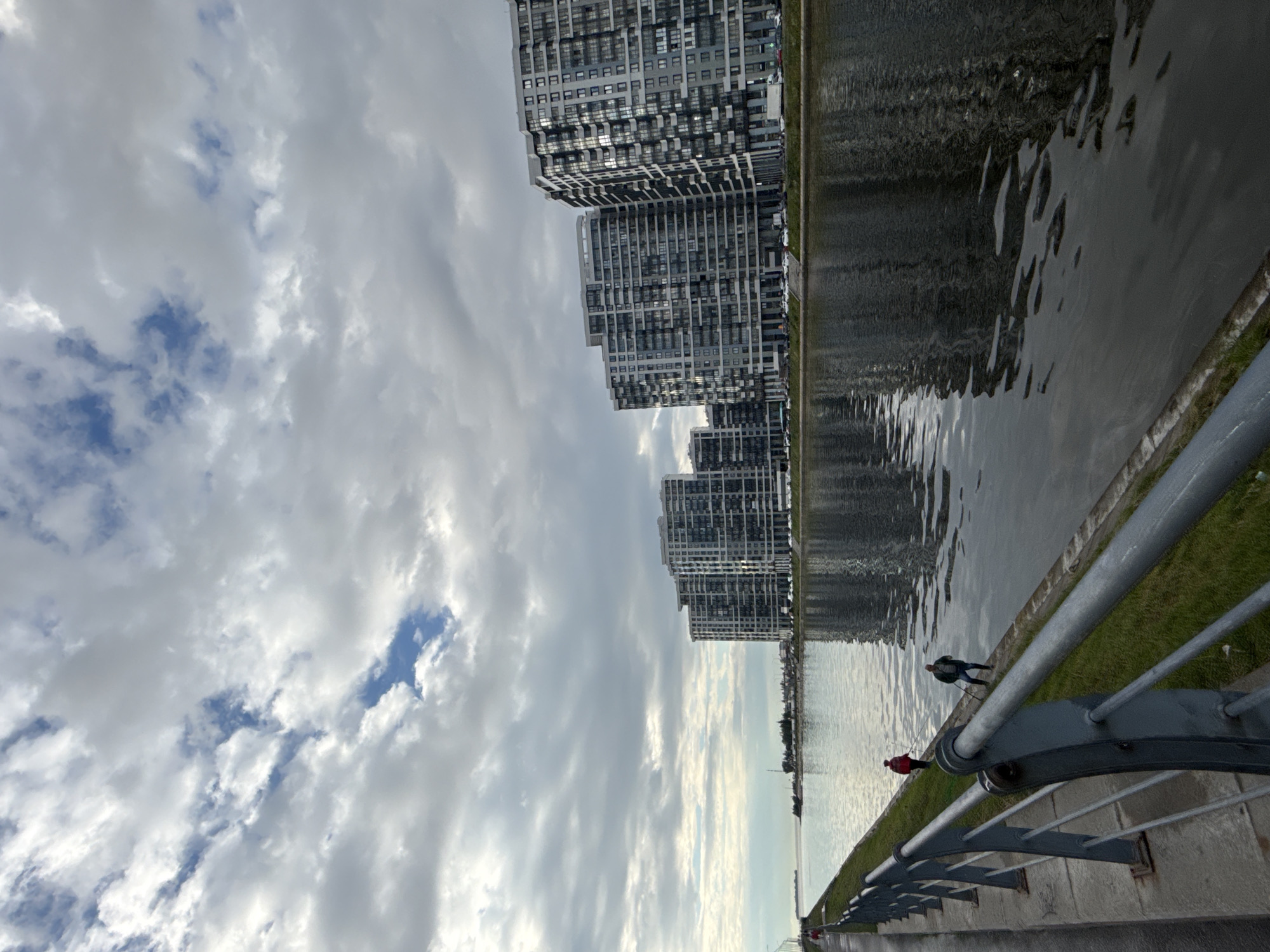 The image depicts a waterfront scene in an urban environment. On the right side of the frame, several large, modern residential buildings with many windows stand parallel to a body of water. The buildings are dark gray and appear to be multi-story. The water reflects the buildings and the overcast sky. On the left side of the frame, a walkway runs alongside the water, separated by a metal railing. Two individuals are visible on the walkway, one wearing a red jacket and the other in a dark jacket, both appearing to be fishing. The sky is filled with large, white and gray clouds, with patches of blue sky visible. The overall impression is of a cloudy day in a developed urban area with a focus on residential architecture and waterfront amenities.
