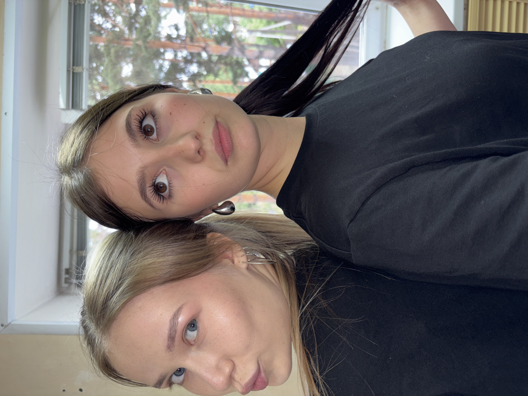 The image shows two young women standing back-to-back, facing away from each other. The woman on the left has blonde hair and is looking over her shoulder with her lips pursed. She is wearing a black t-shirt and hoop earrings. The woman on the right has dark brown hair pulled back into a ponytail and is looking up and to her right. She is wearing a black t-shirt and large, teardrop-shaped earrings. They are indoors, with a window in the background that looks out onto trees. The room appears to be a simple, light-colored space, possibly a classroom or a studio. The specific location in the city of Chaikovsky, Russia, cannot be determined from the visual information provided.
