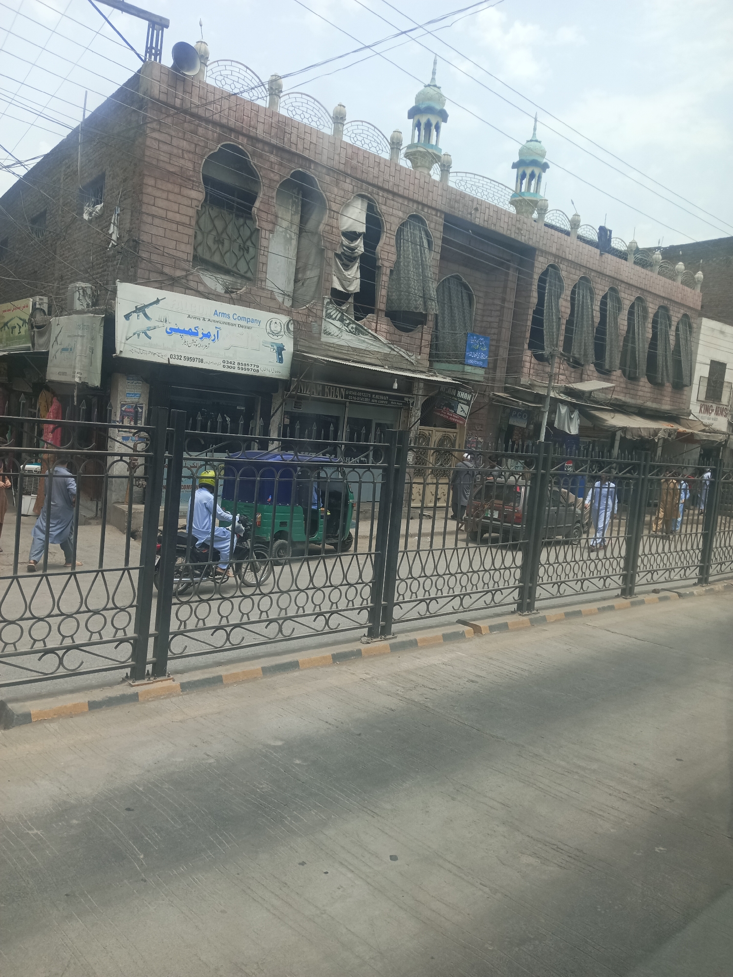 The image shows a street scene in Alam Khan Kalay, Pakistan, with a building that appears to be a mosque or a religious structure with arched windows and minarets. In front of the building, there is a fence. Several people are visible, including a man riding a motorcycle and a person in a tuk-tuk. There are also shops with signage, one of which is for "Arms Company." The overall environment suggests an urban setting with commercial activity.