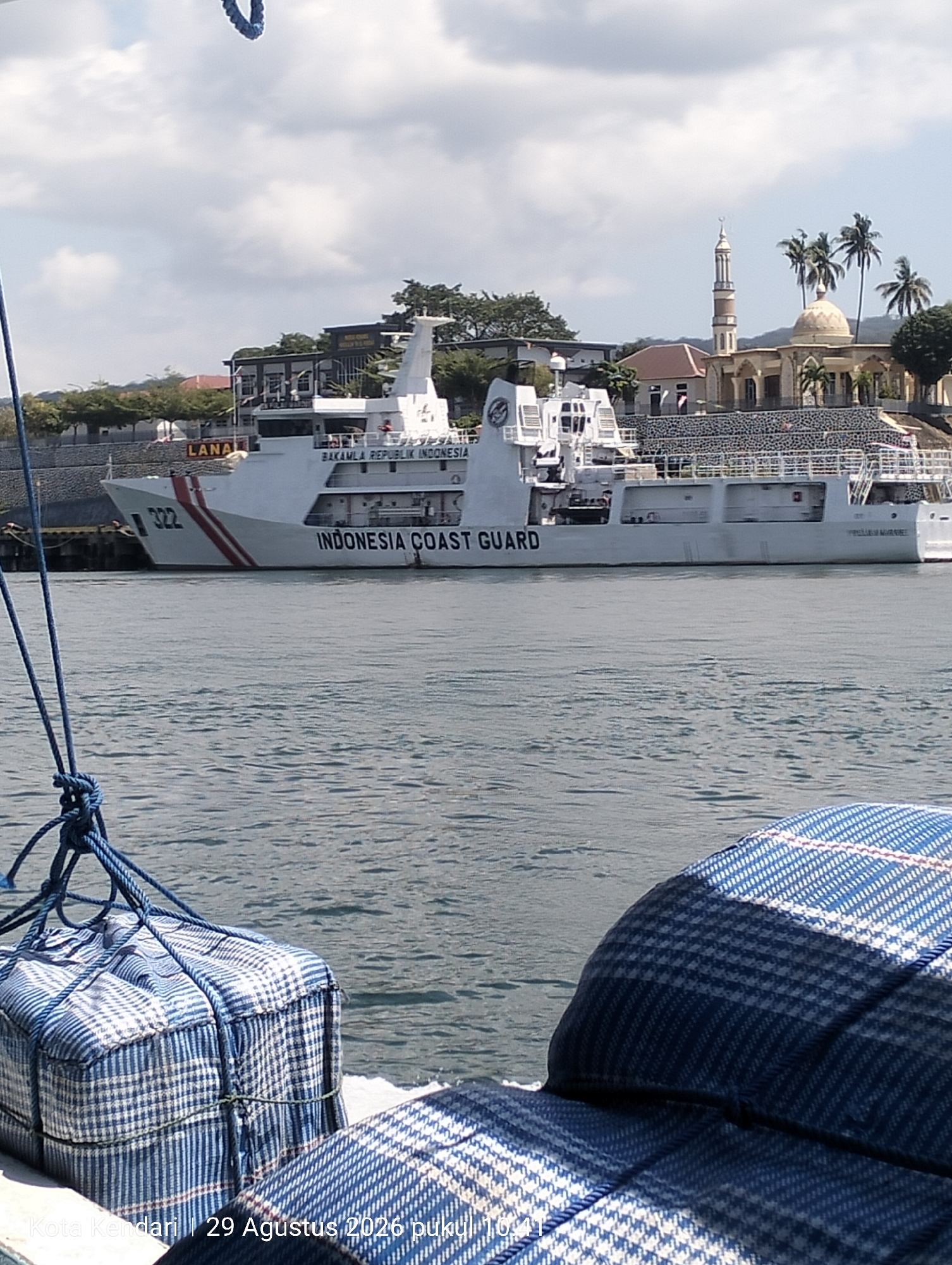 A daylight, medium shot captures a white Indonesian Coast Guard vessel, labeled "BAKAMLA REPUBLIK INDONESIA" and "322" on its hull, docked in the foreground. The vessel is positioned to the left of the frame, with its stern to the right. In the background, a cluster of buildings, including a mosque with a minaret and a dome, is visible. Palm trees dot the landscape behind the buildings. The water in front of the vessel is calm and reflects the sky and surrounding structures. In the foreground, two blue and white checkered bags are tied with ropes, likely part of cargo on a boat from which the photo was taken. The timestamp "10:41" and location "Kota Kendari | 29 Agustus 2026 pukul" are visible in white text at the bottom of the frame.