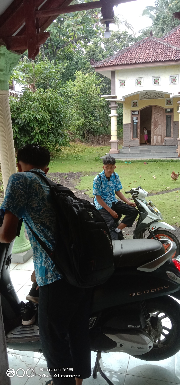 Two young men are seen outside a house. One is sitting on a white scooter while the other stands nearby with a black backpack. The house has brown doors and a red tile roof. A small child is visible in the doorway. Chickens roam on the green lawn in front of the house. A lush green garden surrounds the property.