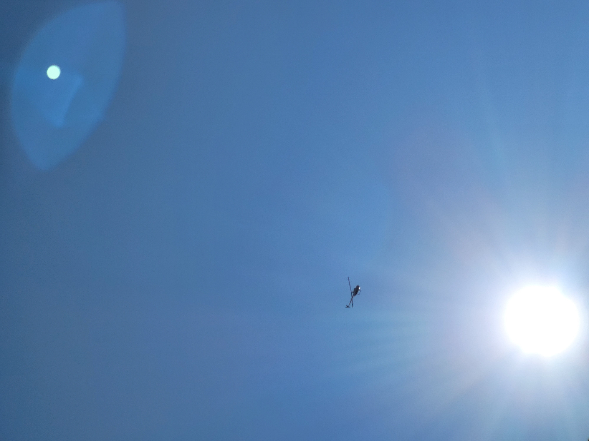 The image is a vertical, low-angle shot of a clear blue sky with a bright sun, creating lens flare. A small, dark silhouette of a helicopter is visible in the middle left of the frame, in flight. The sun is positioned in the lower left, emitting prominent sunbeams. Another, fainter lens flare is present in the upper right quadrant of the sky. There are no people, buildings, or any other recognizable landmarks in the frame, making it impossible to determine its location within the city of Jelgava, Latvia.