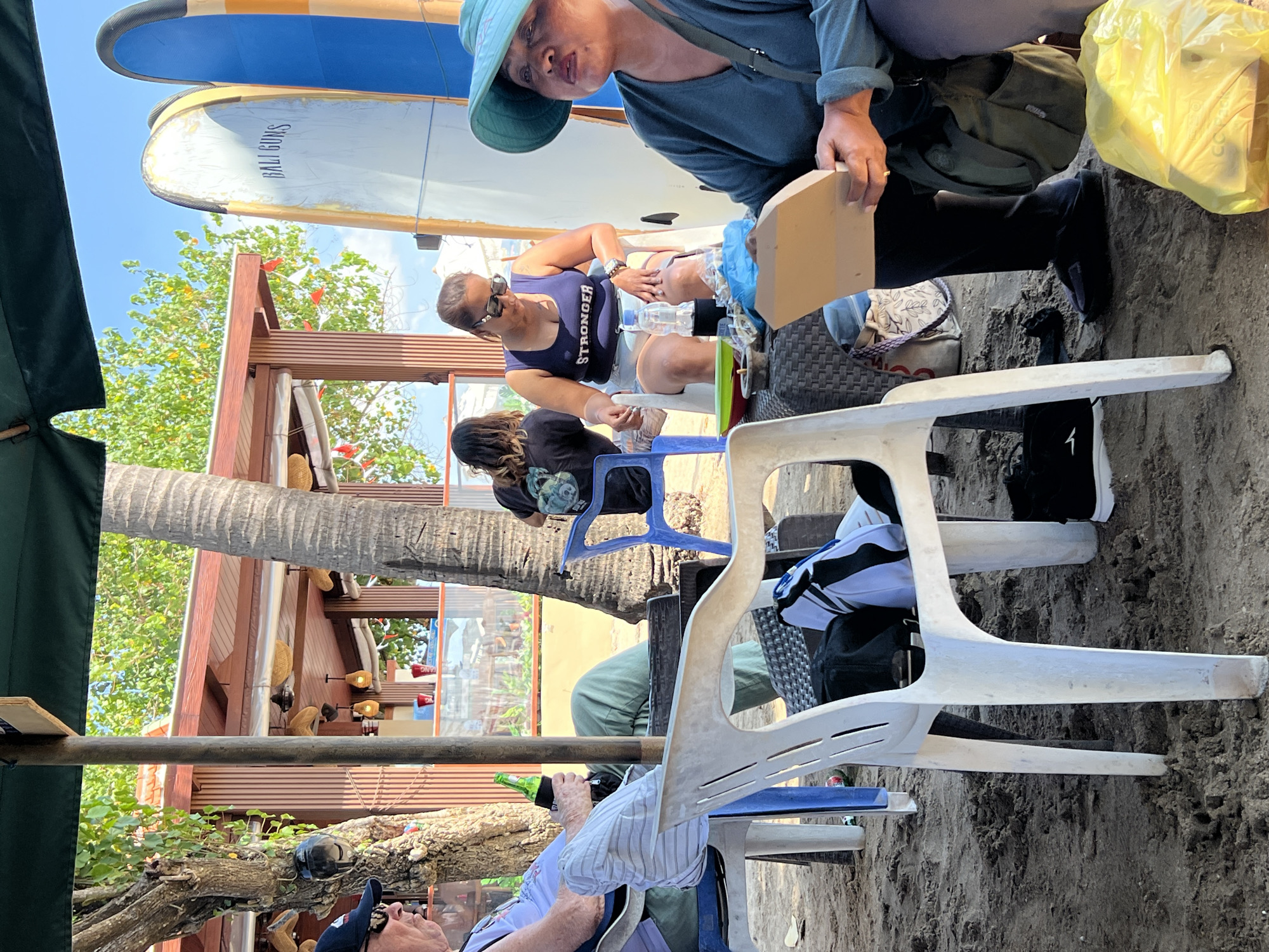 The image depicts an outdoor scene on a sandy beach, likely a cafe or restaurant in Kuta, Indonesia, with a thatched-roof structure in the background. In the foreground, several people are seated on chairs. On the left, a person wearing a cap and sunglasses holds a beer bottle. In the center, a woman in a blue tank top and shorts is seated, with a water bottle and a small plate on a table in front of her. To her right, another woman, wearing a green hat and a teal shirt, is holding a cardboard object. Two surfboards, one with "BALI GUNS" written on it, are propped up behind the seated individuals. Palm trees and lush greenery are visible behind the structure, and the sky is clear and blue. The ground is covered in sand, with some debris and beach chairs scattered around.