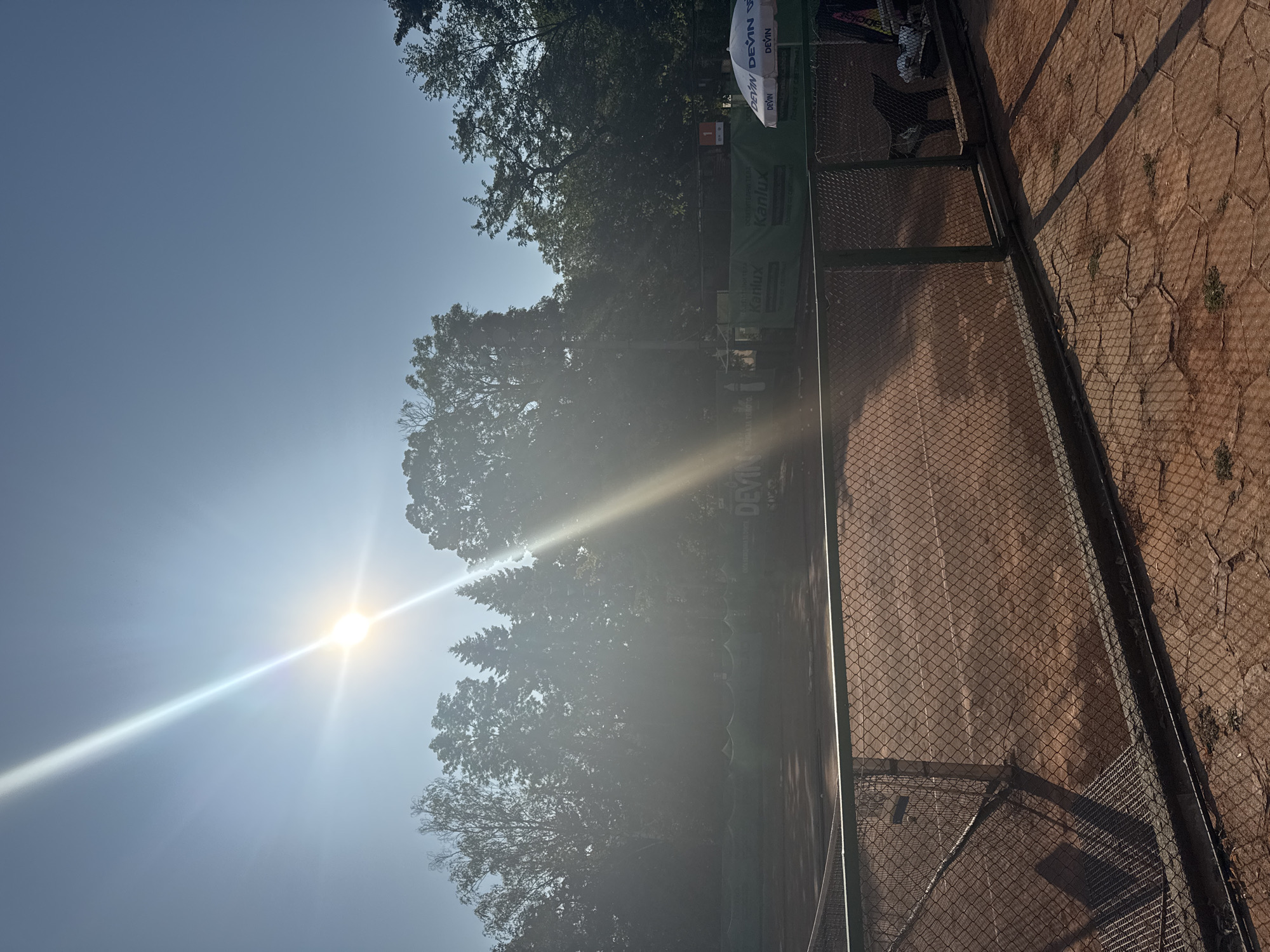 The image shows a sun-drenched outdoor tennis court. The court is made of red clay, with white lines marking the boundaries. A chain-link fence surrounds the court. In the background, trees create a silhouette against the bright sky, and a large sun glare emanates from the upper left corner. A white umbrella with the "DEVIN" logo is visible to the right, as are two chairs. The scene appears to be in a park or sports complex within Sofia, Bulgaria, suggested by the presence of the "DEVIN" brand, a prominent Bulgarian mineral water company.