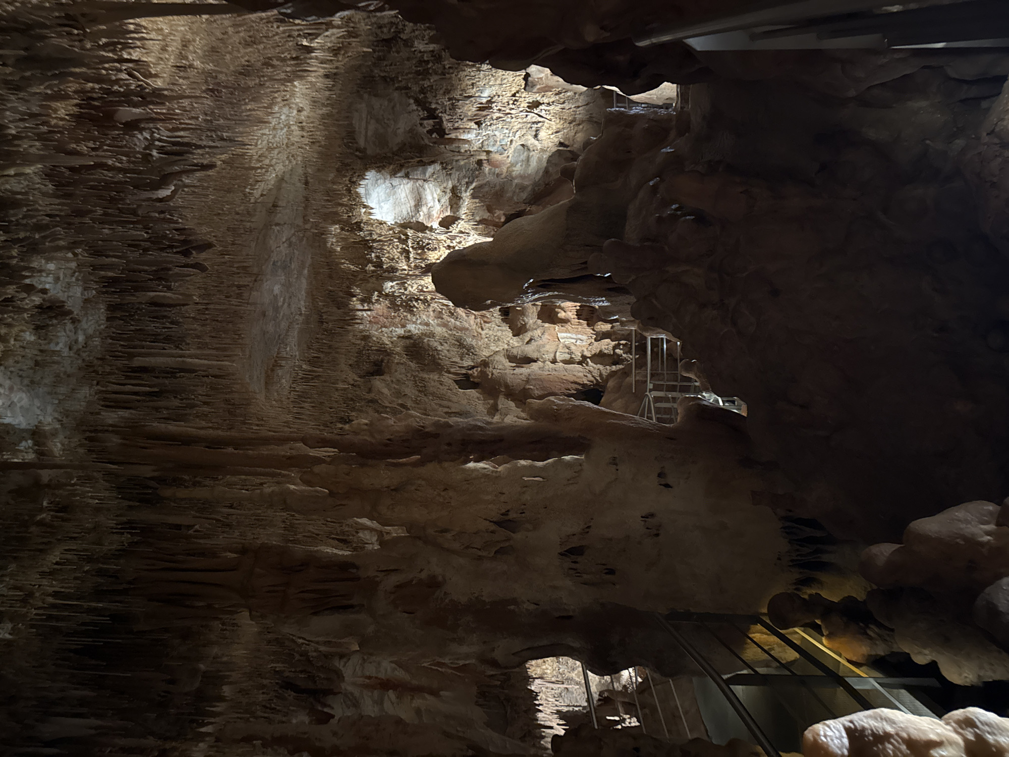 The image depicts the interior of the Cave of Petralona, located in Πετράλωνα, Greece. The cave walls are composed of rough, brown, and tan rock formations, including stalactites hanging from the ceiling on the left side of the image. Metal staircases are built throughout the cave for visitors to navigate the terrain. The lighting is dim and accentuates the contours of the rock formations.