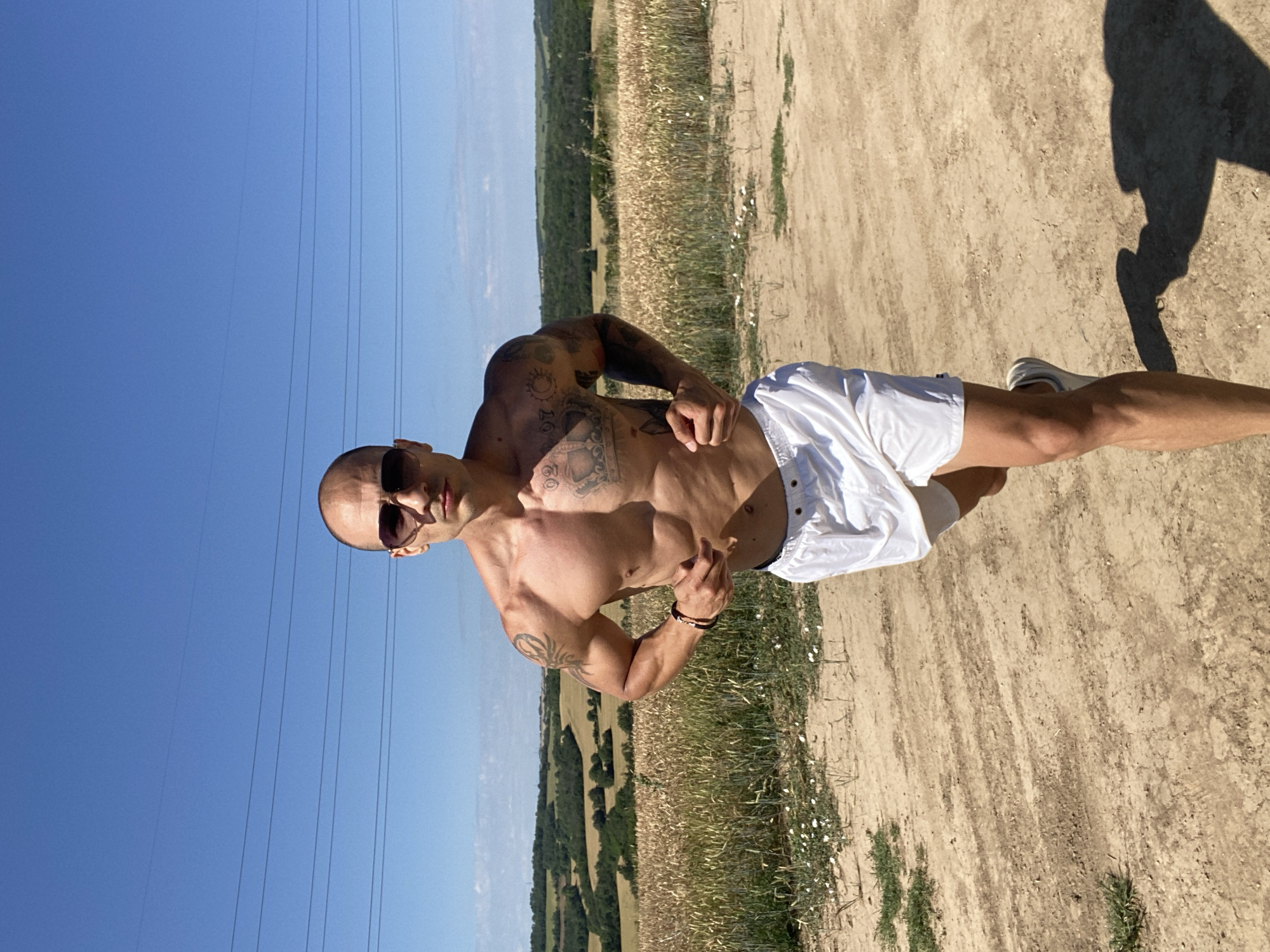 An athletic bald man with sunglasses and visible tattoos on his torso and arms runs on a dirt path. He is wearing white shorts and white athletic shoes. The background consists of a field of dry grass and vegetation, rolling hills with trees, and a bright blue sky with power lines running across it. The scene is set in a rural area, likely a field or countryside, during the day under clear skies.