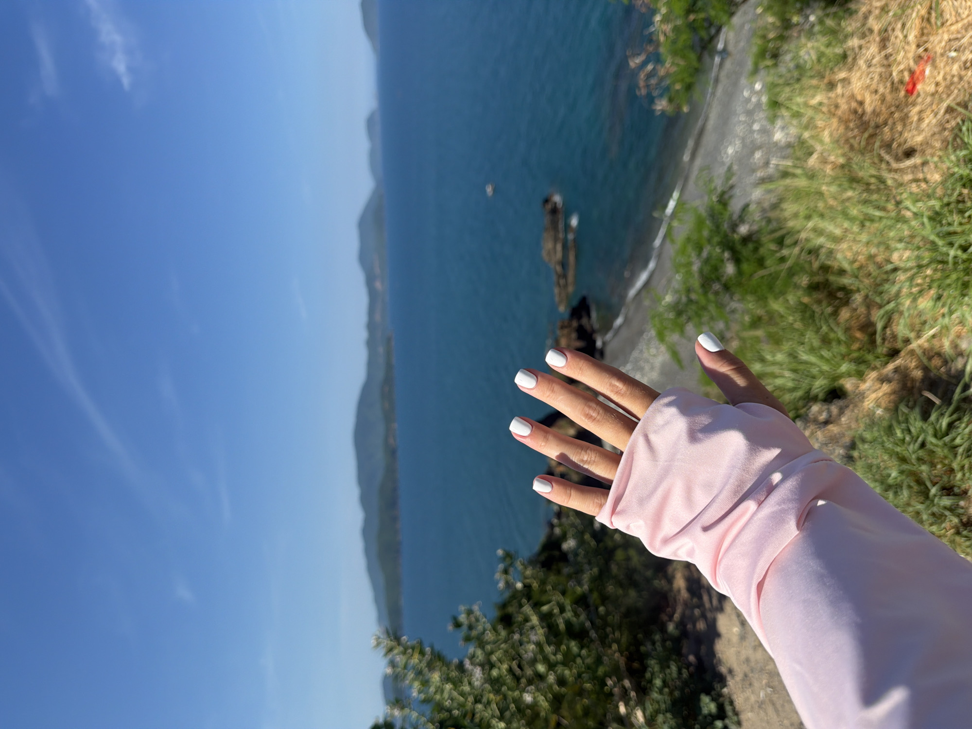 A hand with white nail polish is held up in the foreground, obscuring part of the background. The hand is covered by a pink sleeve, likely part of a shirt or arm warmer. The background shows a scenic view of the ocean with islands in the distance under a clear blue sky with faint clouds. A rocky shoreline with some vegetation is visible below the hand. The location is identified as Nam Nha Trang Ward, Vietnam, suggesting a coastal area. The scene appears to be a snapshot of a person enjoying a view.