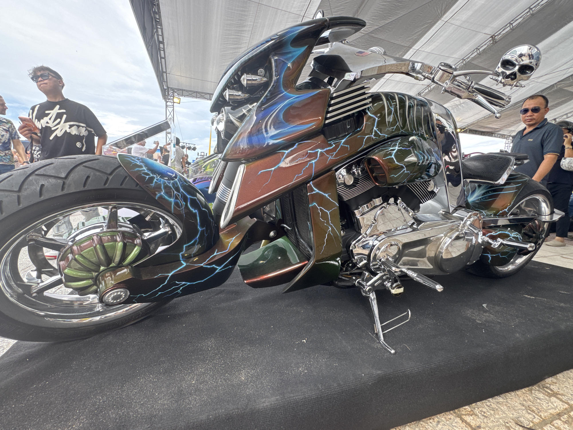 Captured at an undisclosed location, the image showcases a custom motorcycle exhibiting elaborate airbrushing, dominated by a black and brown color scheme accented with blue lightning bolt patterns. The motorcycle, positioned on a black platform, is adorned with chrome features, including a skull-shaped rearview mirror. In the background, two individuals are visible: one in a black t-shirt with white lettering and sunglasses, and another wearing a striped polo shirt with sunglasses. Additional onlookers are present further in the background. The scene is set under a tent-like structure, hinting at an outdoor event or exhibition.