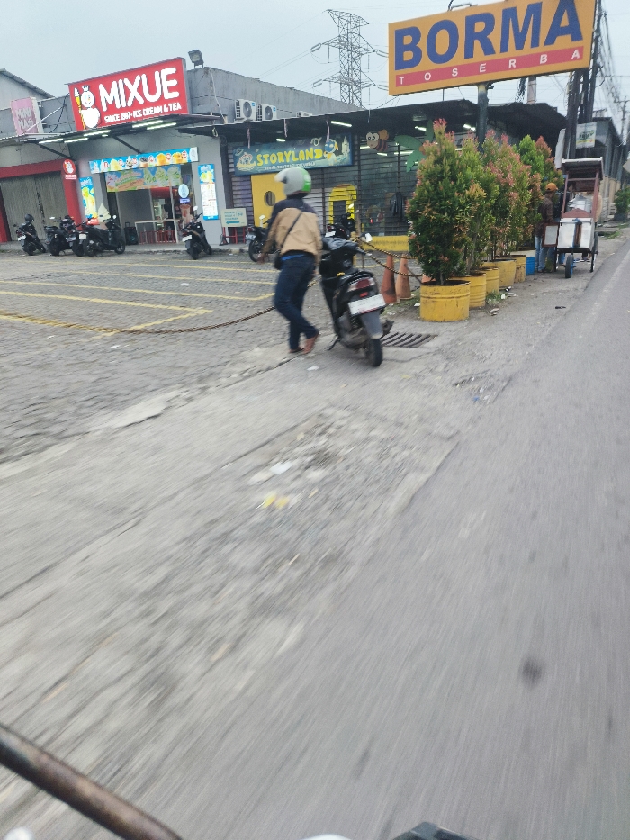The image shows a man in a helmet pushing a black motorcycle on a paved road in Cileunyi, Indonesia (latitude: -6.937, longitude: 107.73). The man is wearing a light blue shirt and dark pants. The motorcycle is facing the camera, with the front wheel off the ground. There is a row of potted plants on the right side of the road and a sign with the words "BORMA TOSERBA" on the left. Several parked motorcycles are visible in the background. There is a shop with a "MIXUE" sign on the right side of the road and an empty parking lot to the left. The scene is taken from a low angle, with the camera facing the road. The image has a slight motion blur effect, likely caused by the camera being moved during the shot. The image is taken during the day, with clear skies.
