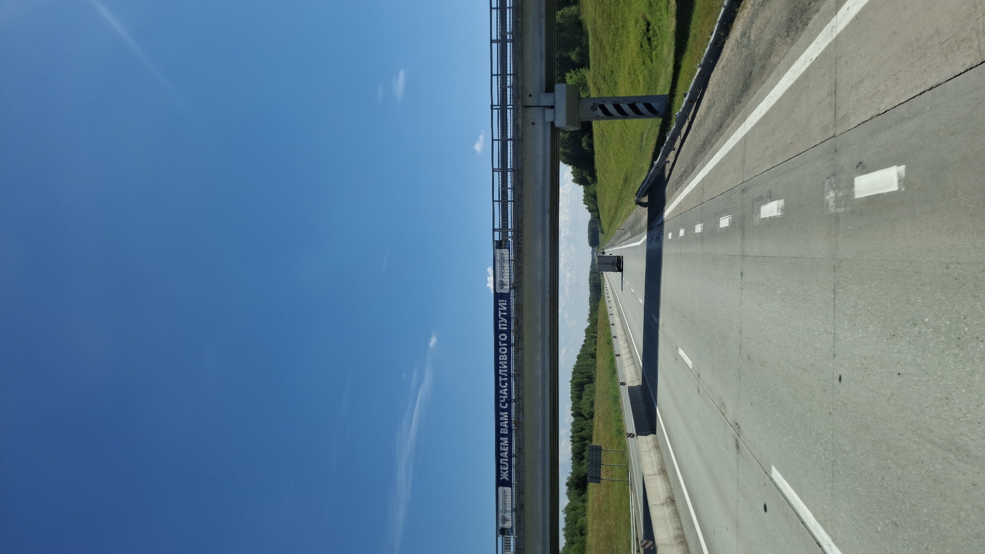The image shows a view from the driver's perspective on a highway in the city of Sokur, Russia. The highway is paved with asphalt and has multiple lanes separated by white lines. There are concrete barriers on the sides of the road. A pedestrian overpass spans the highway, with a banner on it that reads "ЖЕЛАЕМ ВАМ СЧАСТЛИВОГО ПУТИ!" which translates to "We wish you a happy journey!". The sky is clear blue with a few wispy clouds. The surrounding environment consists of green fields and trees. A truck is visible on the highway in the distance. The overall scene depicts a typical highway landscape on a sunny day in Russia.
