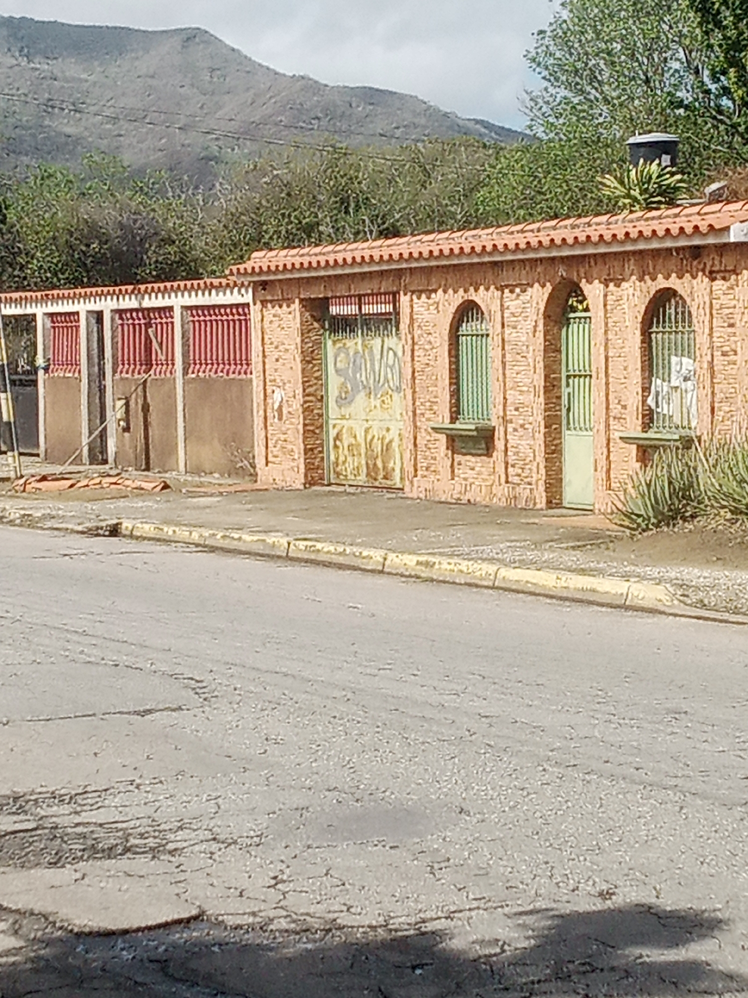 The image shows a street in front of a building with an orange tiled roof and brick walls. The building has several arched openings with green metal grates. Some of the openings have green metal doors, while others appear to be windows. There is graffiti on some of the metal surfaces. To the left of the building, there is a section of red metal fencing. In the background, there are trees and a mountain. The street in the foreground is paved and appears to be cracked and worn. There is a yellow curb along the side of the street. The scene is captured on a clear day, with sunlight illuminating the building and street. This location is in the city of Santa Ana, Venezuela.