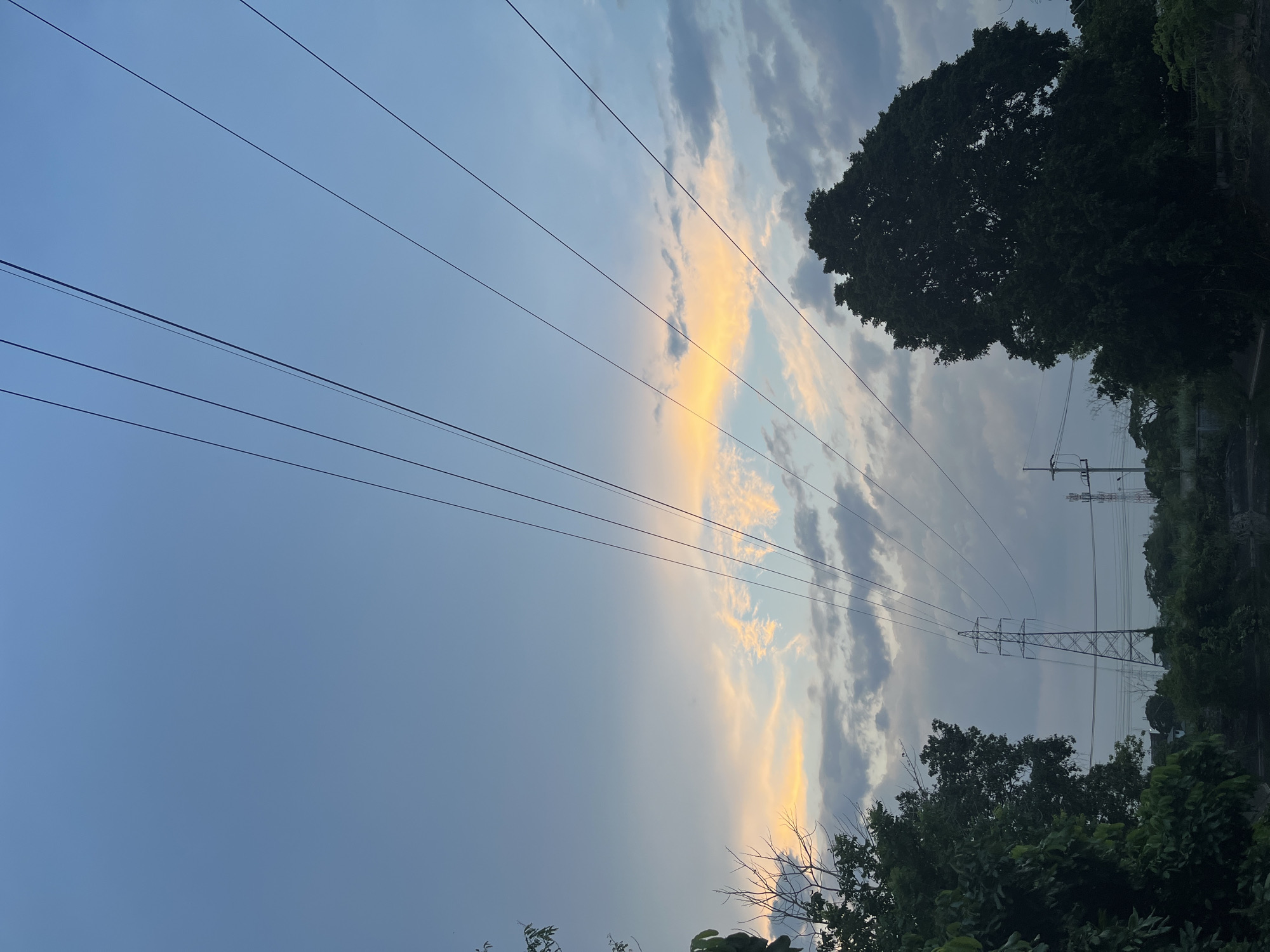 The image depicts a view looking upwards towards a sky with clouds and power lines. A patch of sunset-colored clouds is visible amidst the predominantly blue and gray sky. Dark green trees are present along the bottom and right edges of the frame. In the distance, a transmission tower is visible among the trees. This location is within the city of บ้านกาญจน์กนก 23, Thailand. There are no people visible.